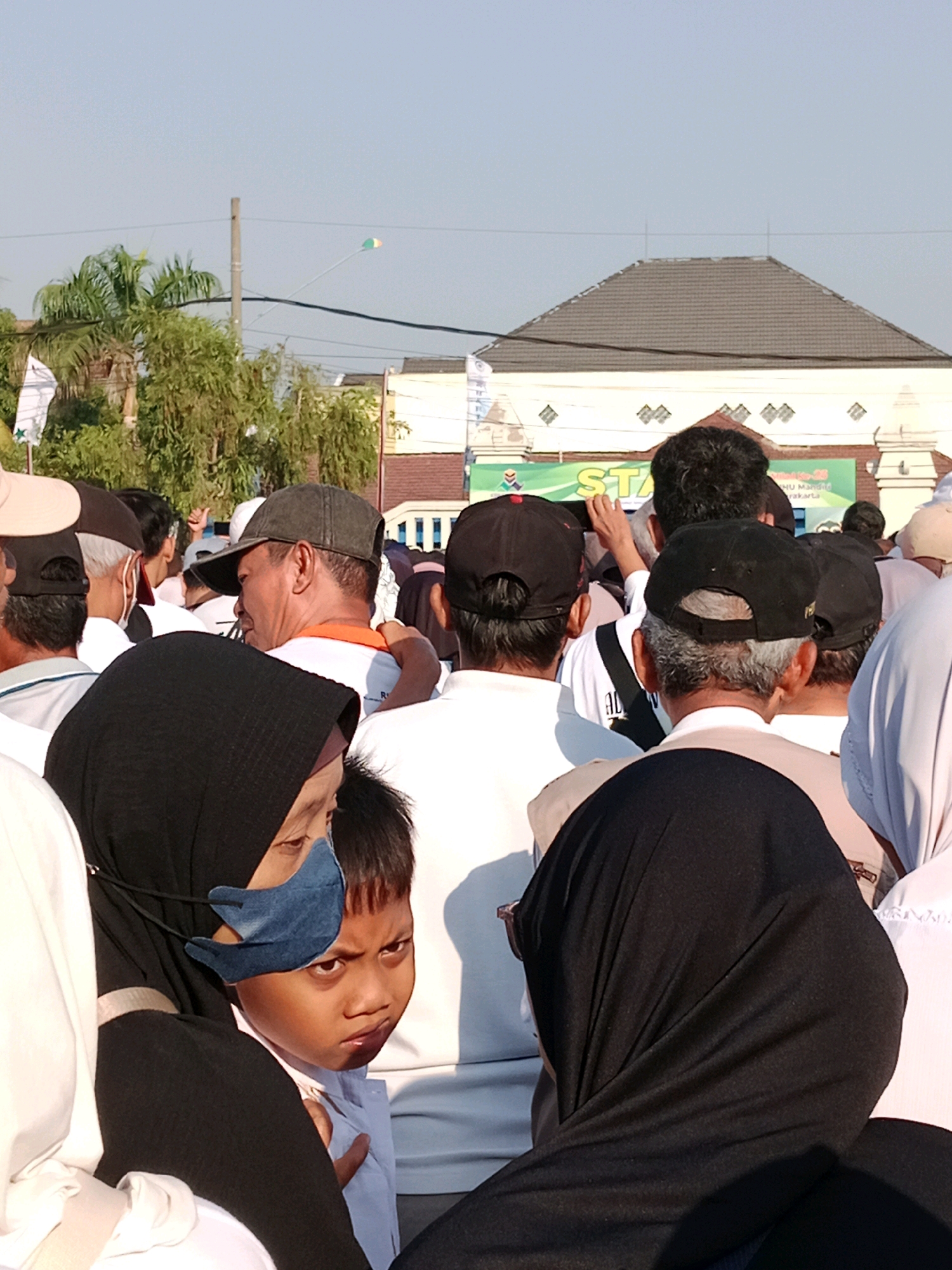A low-angle, outdoor, medium shot captures a crowd of people in what appears to be a public gathering in Surakarta, Indonesia, possibly for an event or ceremony. The sky is a pale blue with no clouds. A tall, slender utility pole with wires stretches across the upper portion of the image. In the distance, a dark, pitched roof of a building is visible, adorned with a decorative pattern of diamond shapes. A green banner with text, partially obscured by the crowd, is also visible in the background.
The foreground is dominated by the backs of people. A woman in the lower left wears a black hijab and a blue face mask. Next to her, a young boy, also wearing a light-colored shirt and a blue face mask, looks towards the viewer with a serious expression. To the right of the boy, a person in a black head covering is prominently featured.
Further back, several individuals are visible. One man wears a dark baseball cap and an orange-trimmed shirt. Another man, facing away from the camera, wears a dark baseball cap. Others in the crowd are dressed in white shirts or casual attire, many wearing face masks. The presence of multiple face masks suggests a context where public health precautions are being observed. The overall atmosphere appears to be one of a crowded event, with people standing and observing.