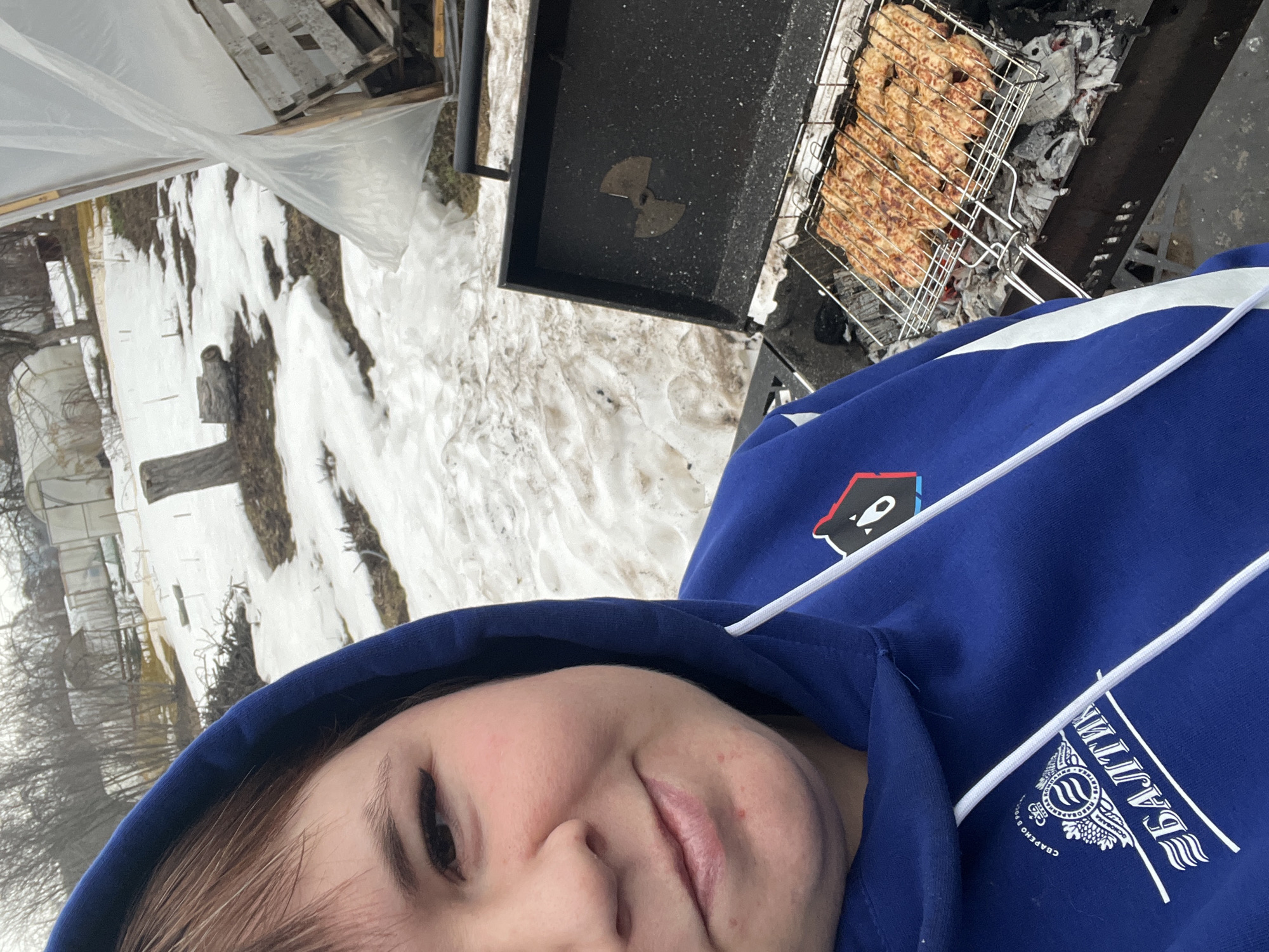 The image is an angled selfie taken in an outdoor environment, likely in a suburban or rural setting. In the foreground, a person wearing a blue hoodie is visible. The hoodie has white drawstrings and a logo on the chest, possibly in Cyrillic. The background features a grill with food cooking over charcoal. Adjacent to the grill is a snow-covered landscape with remnants of winter. The background includes a structure covered in plastic sheeting and bare trees. Based on the Cyrillic logo on the hoodie, and the user's request, it is highly probable that this picture has been taken in the city of Chaikovsky, Russia.