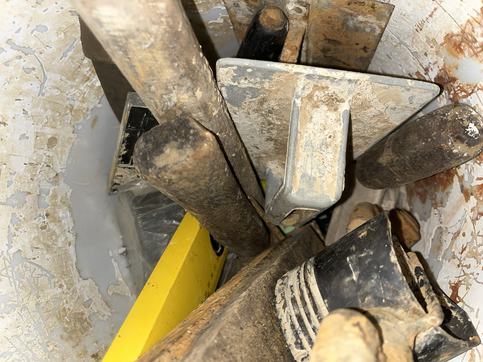The image shows a collection of worn and dirty construction tools inside a white bucket. The tools appear to be made of metal and wood, with some showing signs of rust and age. Among the visible tools are trowels, a spirit level, and what looks like a handle or shaft from another tool. The setting is an indoor or covered area, judging by the lighting and the condition of the bucket's interior, which is stained with what appears to be old plaster or cement. The ground or surface around the bucket is not visible. There are no people or animals present in the image, and no specific activity is depicted, other than the tools being stored together. The notable detail is the general state of disuse and wear on the tools, suggesting they are either old or have been recently used in a demanding task. No visible text is present on the tools or the bucket. The overall impression is one of a work-in-progress or a tool storage area in a construction or renovation environment.