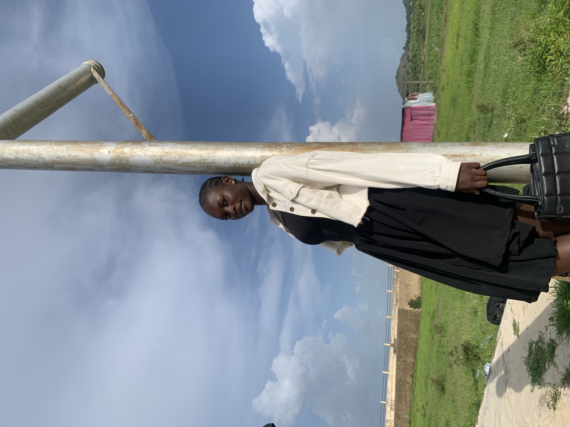 A young Black woman stands leaning against a rusty metal pole in Gwafan, Nigeria. She is wearing a black dress and a cream-colored jacket, holding a black handbag. The background shows a grassy field with a brick wall and a small pink building. The sky is cloudy.