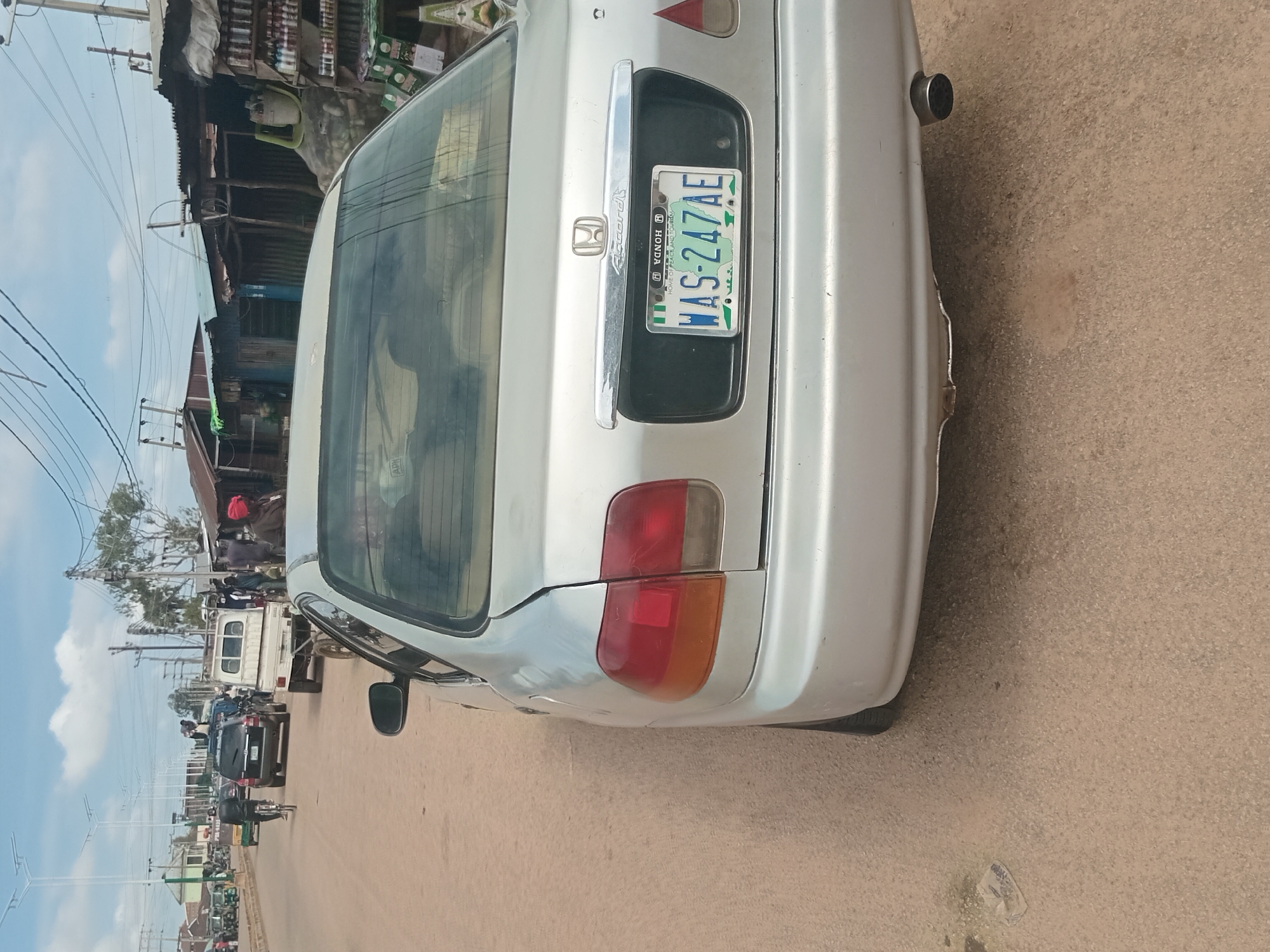 The image shows a silver Honda Accord sedan parked on the side of a dusty, unpaved road. The car's rear end is prominently featured, with its license plate clearly visible. The license plate reads "WAS-247AE" and appears to be a Nigerian plate, indicated by the green and white design and the text "HOME OF PEACE AND TOURISM." In the background, there are various structures, including makeshift stalls with corrugated metal roofs, suggesting a market or roadside commercial area. Several other vehicles, including a white pickup truck, a black SUV, and what appear to be motorcycles and tricycles, are present on the road, indicating activity and traffic. People are also visible in the background, some walking and others near the stalls. The overall environment suggests a bustling, perhaps informal, urban or semi-urban setting. The sky is partly cloudy. The location is identified as Bukuru, Nigeria.