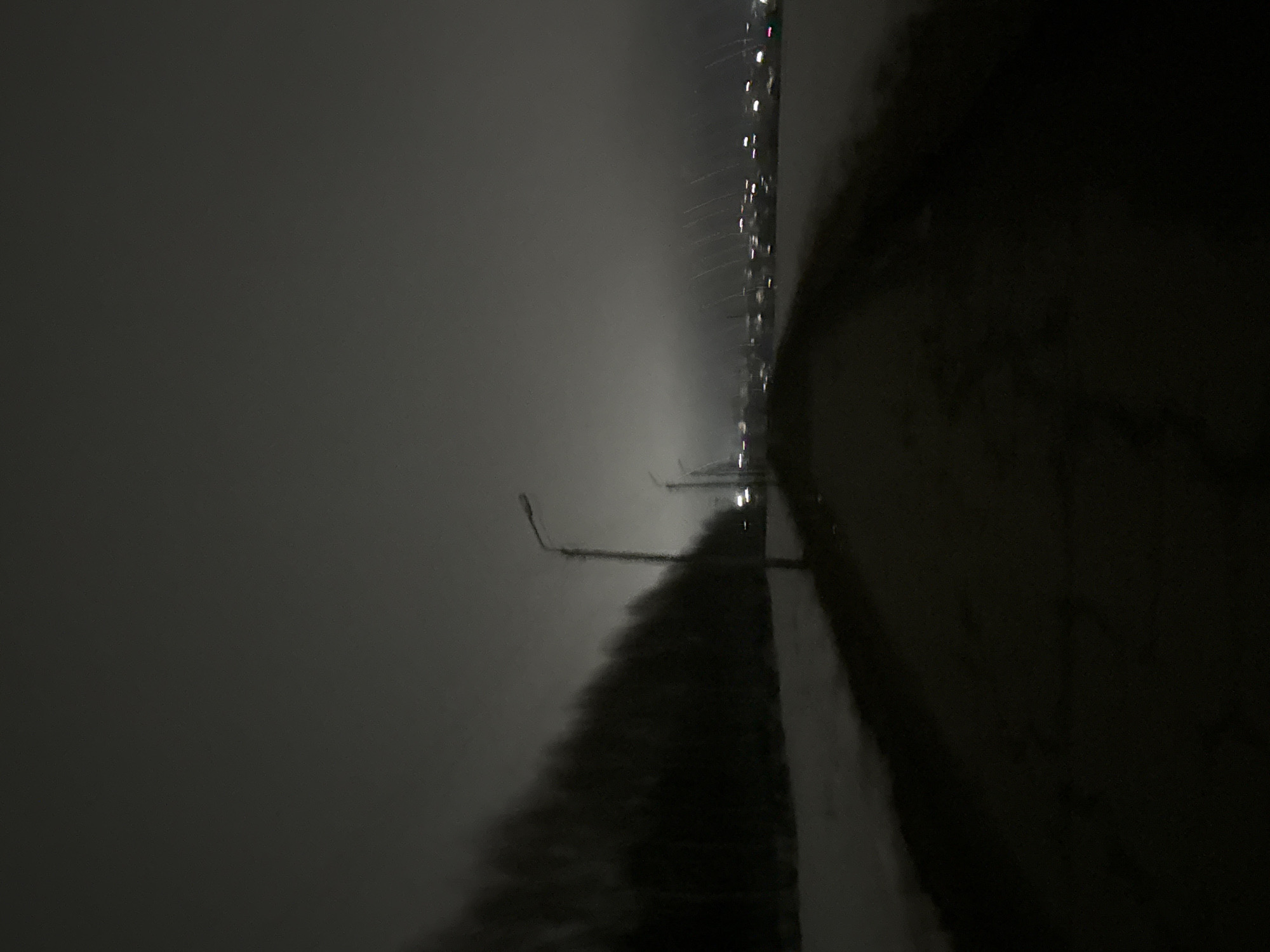 The image shows an outdoor scene at night. A street or road is visible, flanked by what appears to be a line of trees or bushes on one side and a series of streetlights on the other. The overall lighting is dim and murky, suggesting a foggy or overcast condition. The scene appears to be in Фоки, Russia, given the instructions.