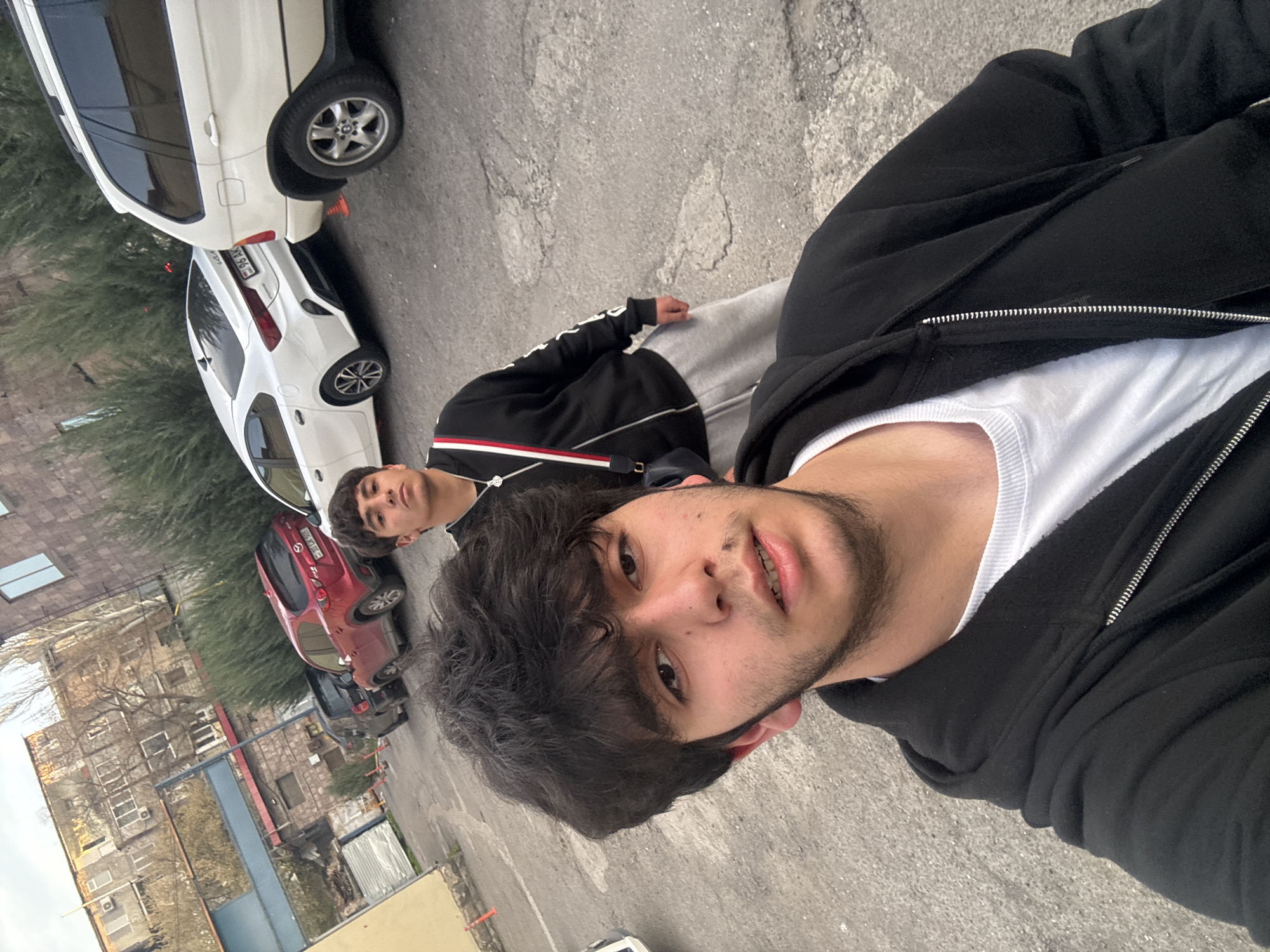 The image shows two young men on a street in Yerevan, Armenia. The man closest to the camera is wearing a black zip-up hoodie over a white t-shirt. He has dark hair and a trimmed beard. The second man, further away, is wearing a black jacket with a red and white stripe. They are standing on a cracked gray asphalt road. In the background, there are several cars parked along the street, including a red Mazda, a white Toyota, and a white SUV. There is also a multi-story building with air conditioning units visible. The sky is overcast, and the buildings appear to be older construction.
