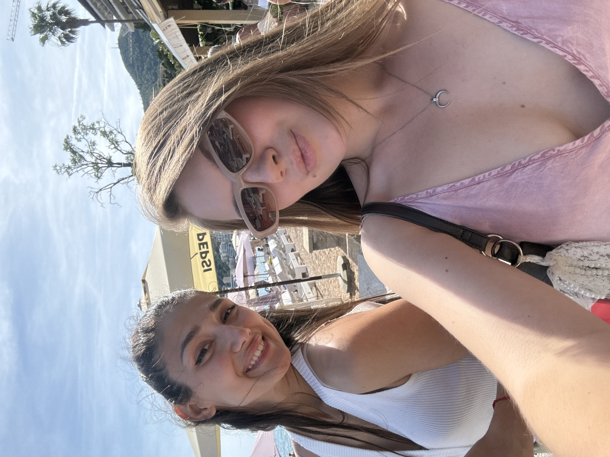 This image captures two young adult females taking a selfie on a sunny day at a beach in Budva, Montenegro. The female on the left has dark brown hair, partially pulled back, and is smiling directly at the camera with her teeth visible. She is wearing a white ribbed tank top and a red bracelet on her left wrist. The female on the right, taking the photograph, has long, lighter brown hair and is wearing light-colored rectangular sunglasses. A small mole is visible above her upper lip on the right side. She is wearing a pink garment with a deep V-neck and a silver chain necklace with a crescent moon pendant. A black bag strap with a white lace and pearl tassel is visible over her left shoulder. The background features a sandy beach with numerous white sun loungers and various beach umbrellas, including pink ones and a prominent yellow umbrella displaying the reversed text "PEPSI". The Adriatic Sea is visible on the far left. Behind the beach, coastal buildings are visible, situated at the base of lush green mountains. A large tree with sparse foliage and a tall palm tree are present, along with a construction crane visible against the blue sky in the upper right. The sky is clear with minimal scattered clouds.