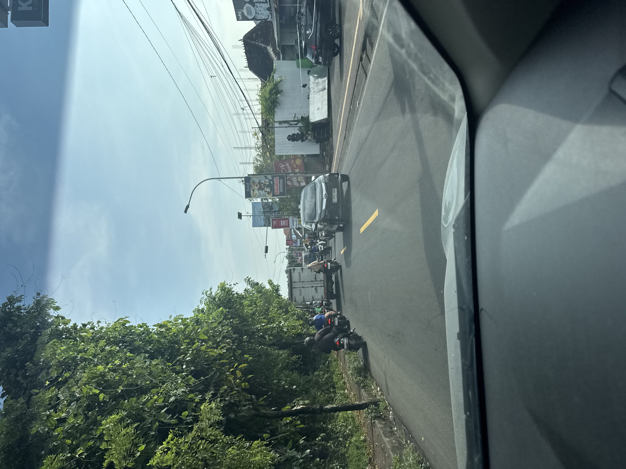 Captured from inside a vehicle, the image presents an eastward view of a roadway in Jimbaran, Indonesia. The road, marked with yellow lines, accommodates several vehicles, including a dark-colored sedan directly ahead and a white truck further in the distance. Motorbikes, with riders wearing helmets, are seen weaving through traffic. Buildings and billboards line the sides of the road, with lush, green trees and foliage bordering the road's edge. Overhead, power lines extend across the sky. The sky is bright, indicating daytime. The vehicle's dashboard and a portion of the side window are visible in the foreground.