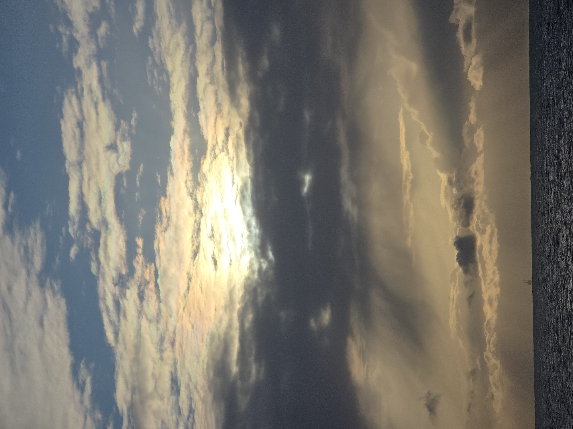 The image depicts a sky filled with various cloud formations, with the sun shining through a break in the clouds, creating a bright and iridescent effect. To the right of the frame, a strip of water is visible. The location is unknown.