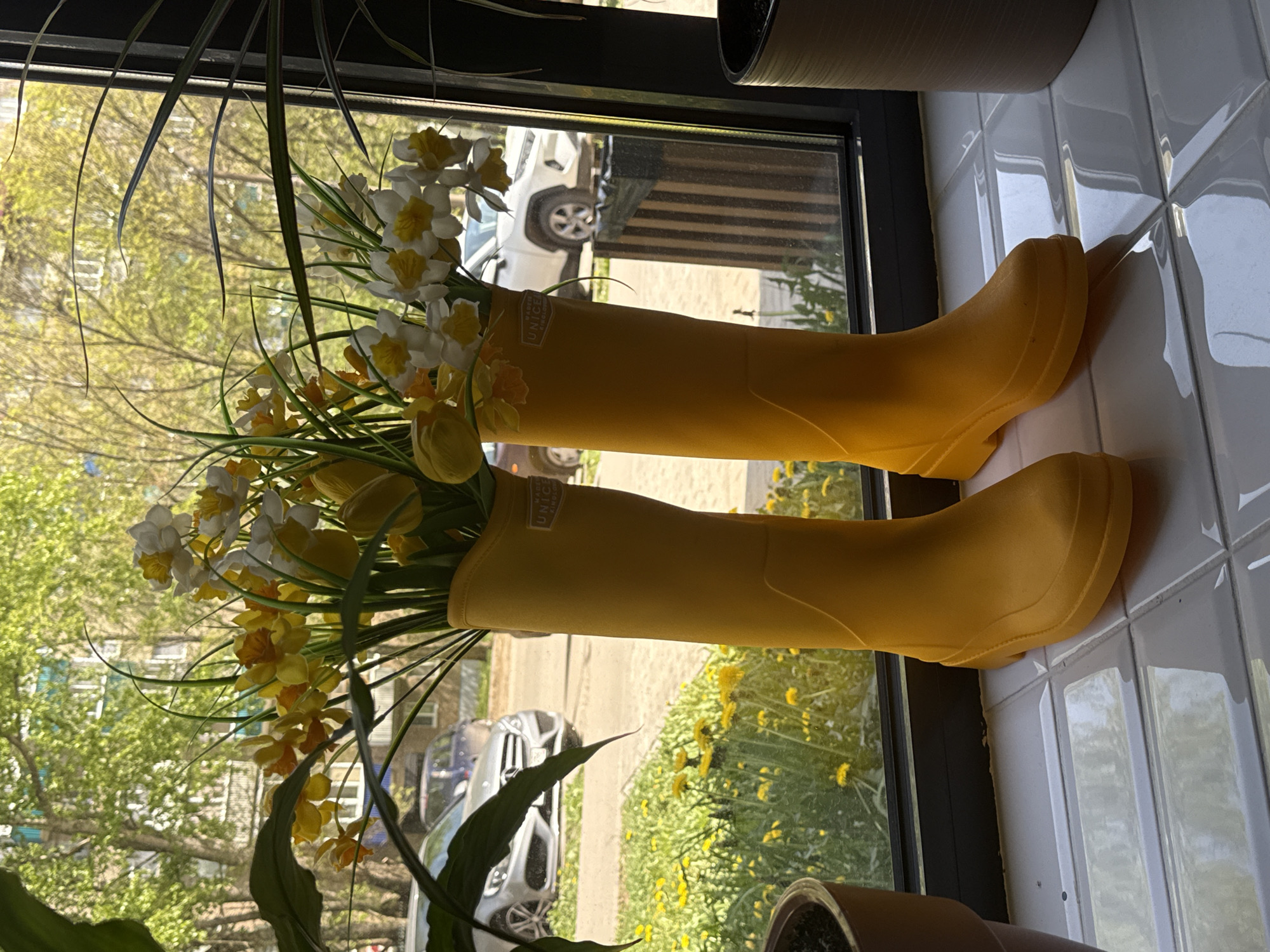 Two yellow rain boots filled with flowers are positioned on a window sill. The flowers include daffodils and tulips. Outside the window, a street scene with cars and yellow dandelions is visible. The location is Borok, Russia. The window sill is covered in white ceramic tiles.