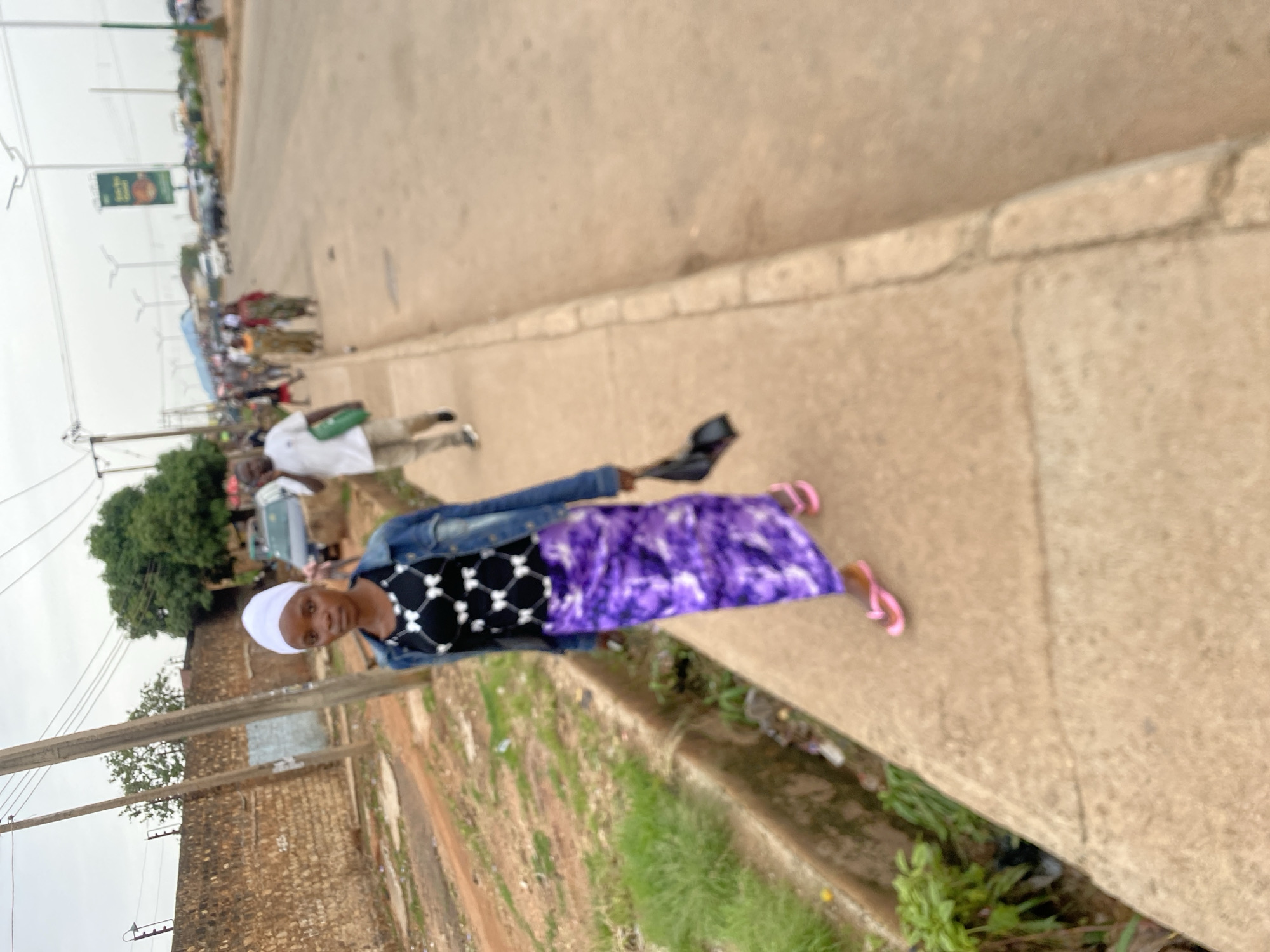 A young woman walks down a sidewalk in Bukuru, Nigeria, holding a black bag. She is wearing a white head covering, a black and white patterned top, a long purple skirt, and pink flip-flops. The sidewalk is made of concrete and has a drainage ditch running alongside it filled with grass and debris. In the background, there is a street with traffic and other pedestrians. A brick wall is visible on the left side of the image. Overhead wires and streetlights are also present. The sky is overcast.