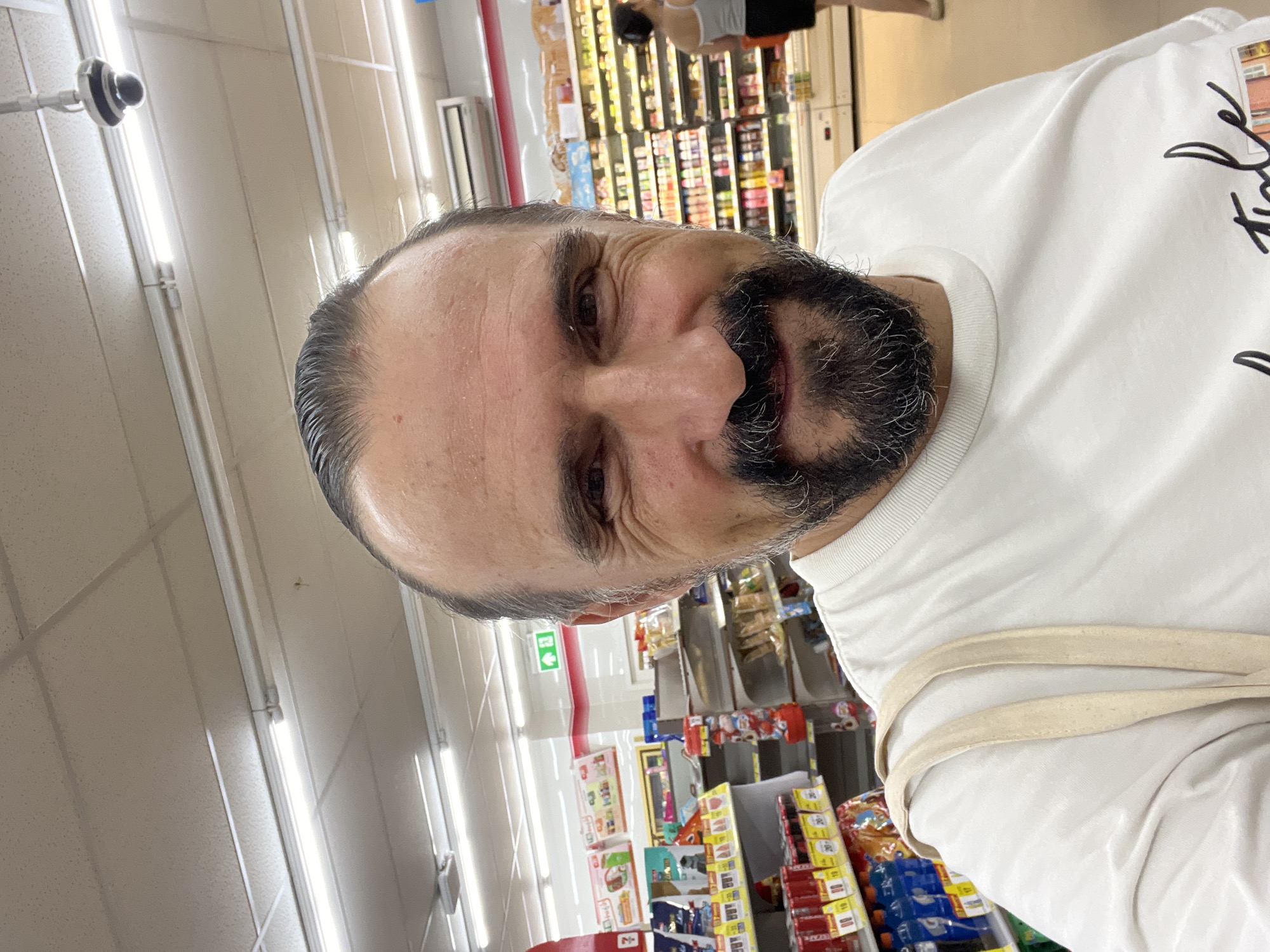 A male individual with dark, slicked-back hair, a graying beard, and a mustache is depicted in a close-up selfie taken within a convenience store in the city of บ้านบางรักษ์, Thailand. He wears a light-colored, possibly cream or off-white, crew-neck t-shirt and has a light-colored canvas tote bag strap over his right shoulder. His expression is a slight smile, with visible crinkles around his eyes. The background reveals a retail environment with white drop ceilings, fluorescent tube lighting, and a black dome security camera mounted on the ceiling in the upper right. An air conditioning unit is visible on the right wall. Numerous product shelves line the store; on the left, shelves hold packaged goods, and on the right, refrigerated display cases contain various beverages and food items. A green exit sign with a white arrow pointing right is positioned above a shelf on the left. In the mid-ground on the right, a female individual with dark hair pulled back, wearing a light-colored tank top and a dark skirt, is observed from behind, seemingly examining products in a refrigerated aisle.