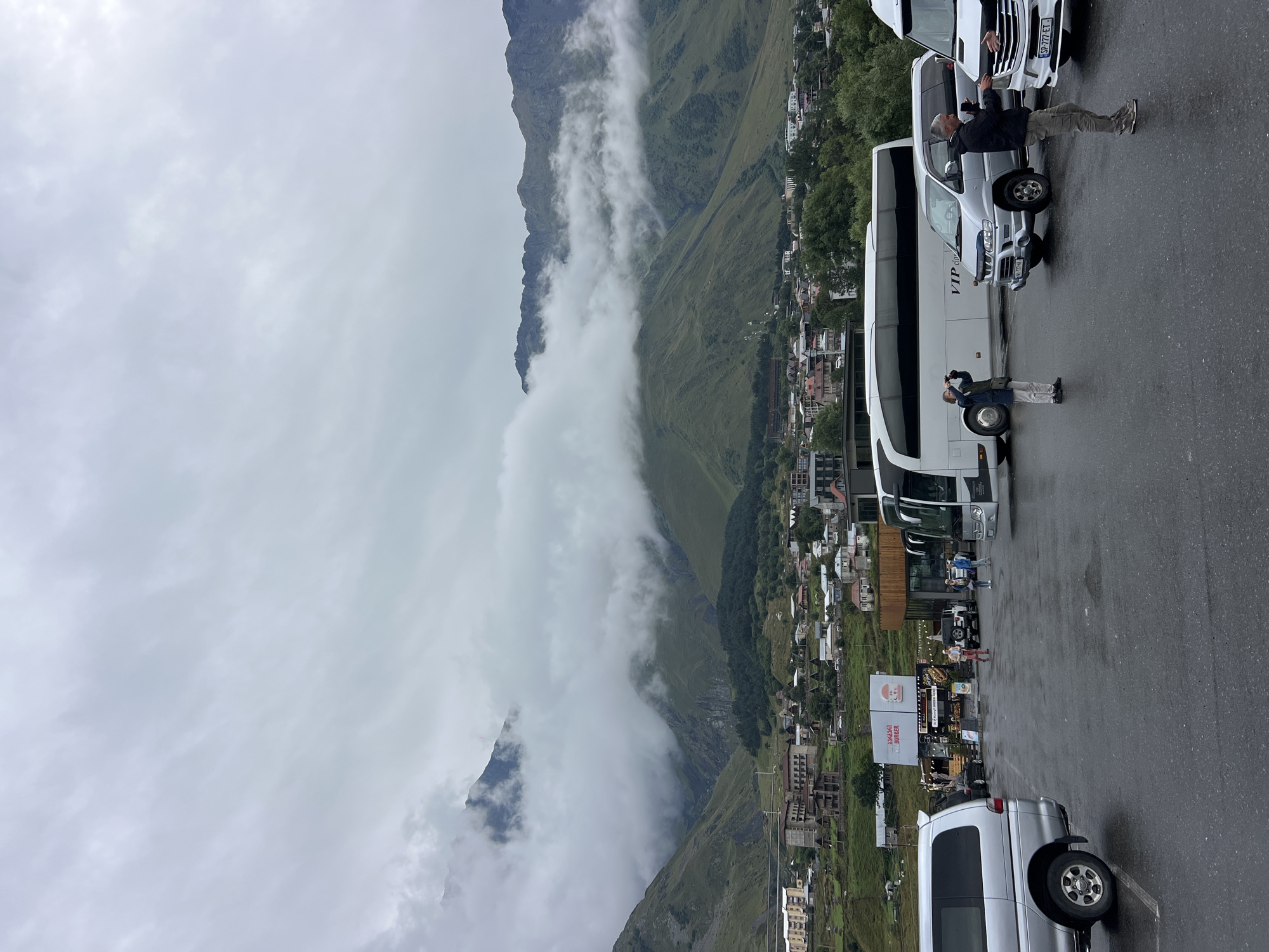In Stepantsminda, Georgia, under a cloudy sky, a parking lot is depicted with several vehicles, including a silver minivan in the foreground on the left, a white bus with "VIP" lettering, and two white vans. People are present in the parking lot, with one person photographing the landscape, another engaged in a conversation with someone partially visible, and others walking near a food establishment with a "Burger" sign. In the background, a town is nestled against lush green mountains, some of which are partially obscured by fog and clouds. The ground of the parking lot appears wet, suggesting recent rain.