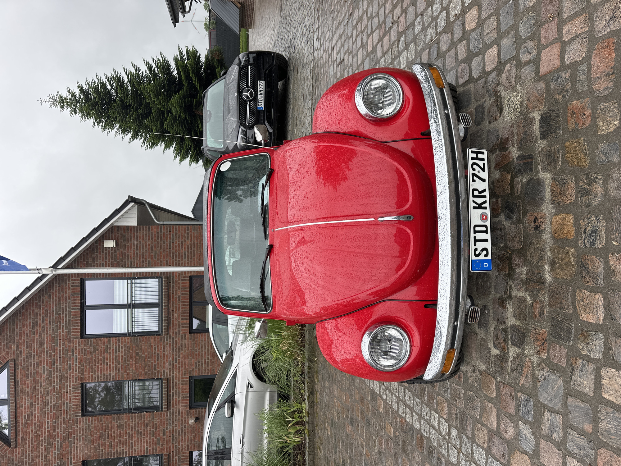 The image depicts an outdoor scene in Deinste, Germany, on an overcast day with visible rain. In the immediate foreground, a bright red, classic Volkswagen Beetle is parked centrally, facing the camera. The car's surface is covered with numerous small raindrops. It features chrome bumpers, circular headlights, and a central chrome strip on the hood. A German license plate, "STD KR 72H," is mounted on the front bumper. To the right and behind the Beetle, a black Mercedes-Benz SUV, also covered in raindrops, is partially visible, showing its front grille with the Mercedes-Benz emblem and a German license plate "STD WH 777". To the left and behind the Beetle, a silver Ford S-MAX is partially visible, showing its front quarter and side profile, alongside green vegetation bordering the paved area.
The ground is composed of a wet, multi-colored cobblestone driveway. The background features a two-story brick house with dark-framed windows, some equipped with external railings and internal blinds. A white flagpole with a partially unfurled blue flag is attached to the house's gable end on the right. A tall, slender evergreen tree stands behind the Mercedes-Benz SUV, its dark green foliage contrasting with the grey, uniformly overcast sky. In the distant background, faint structures resembling wind turbines are discernible through the tree's branches. No individuals are present in the scene.