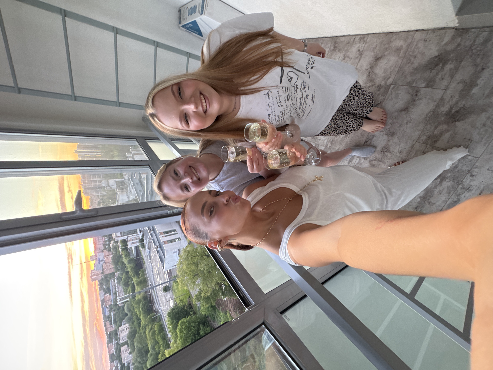Three women are posing for a selfie near a window with a view of Izhevsk, Russia, during sunset. The blonde woman on top wears a white t-shirt with black writing and leopard-print pants. The woman in the middle wears a gray t-shirt. The woman taking the selfie wears a white tank top and a gold necklace. Each woman is holding a glass of champagne. The window reveals a cityscape with trees, buildings, and a colorful sunset. The interior features a grey tiled floor and a white ceiling.