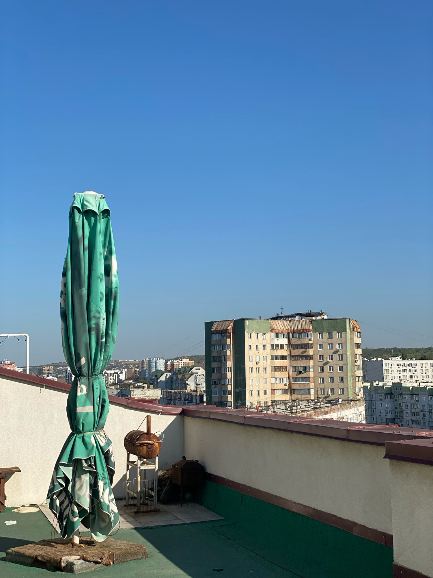 The image depicts a rooftop scene in Chișinău, Moldova, on a clear day. In the foreground, a large, green, closed patio umbrella with a repeating pattern stands vertically. The umbrella is tied in the middle with a string and its fabric is gathered at the base. Adjacent to the umbrella, on a metal stand, is a small, rusty barbecue grill. Further to the right, another dark metal object, possibly a grill or bin, is visible. The rooftop itself is covered with a green surface and bordered by a low, light-colored wall with a brown trim. In the background, a cluster of multi-story apartment buildings dominates the cityscape, with a clear blue sky overhead. The architecture of the buildings suggests a Soviet-era or post-Soviet urban environment typical of Eastern European cities.