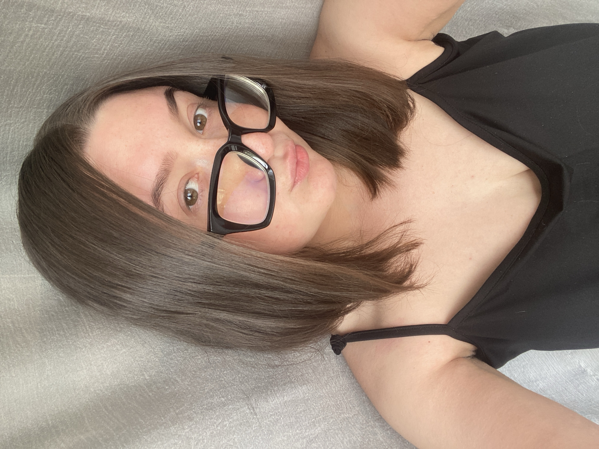 A woman is lying on a gray fabric surface, possibly a bed or couch. She is wearing a black top with spaghetti straps and black-framed glasses. Her hair is straight and brown. The location is unknown, and there is no evidence suggesting Chaikovsky, Russia.