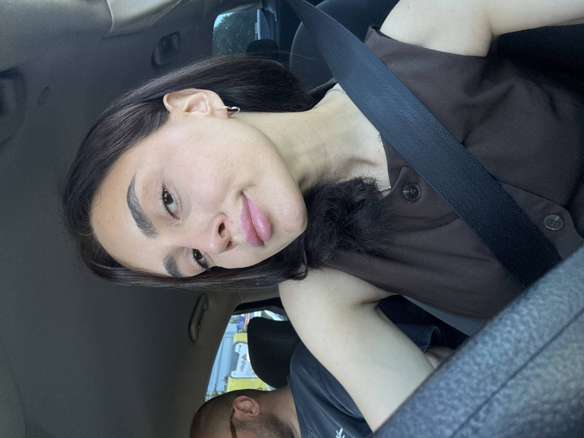 A young woman with dark hair and light skin is in the passenger seat of a car, looking directly at the camera. She is wearing a brown sleeveless shirt with two buttons. A black seatbelt is across her chest. To her left, a man with glasses and a dark shirt is visible. The interior of the car appears to be a standard sedan. Outside the car window, a yellow truck with blue and white signage is visible, suggesting a commercial area. The image is a selfie taken from inside the vehicle.