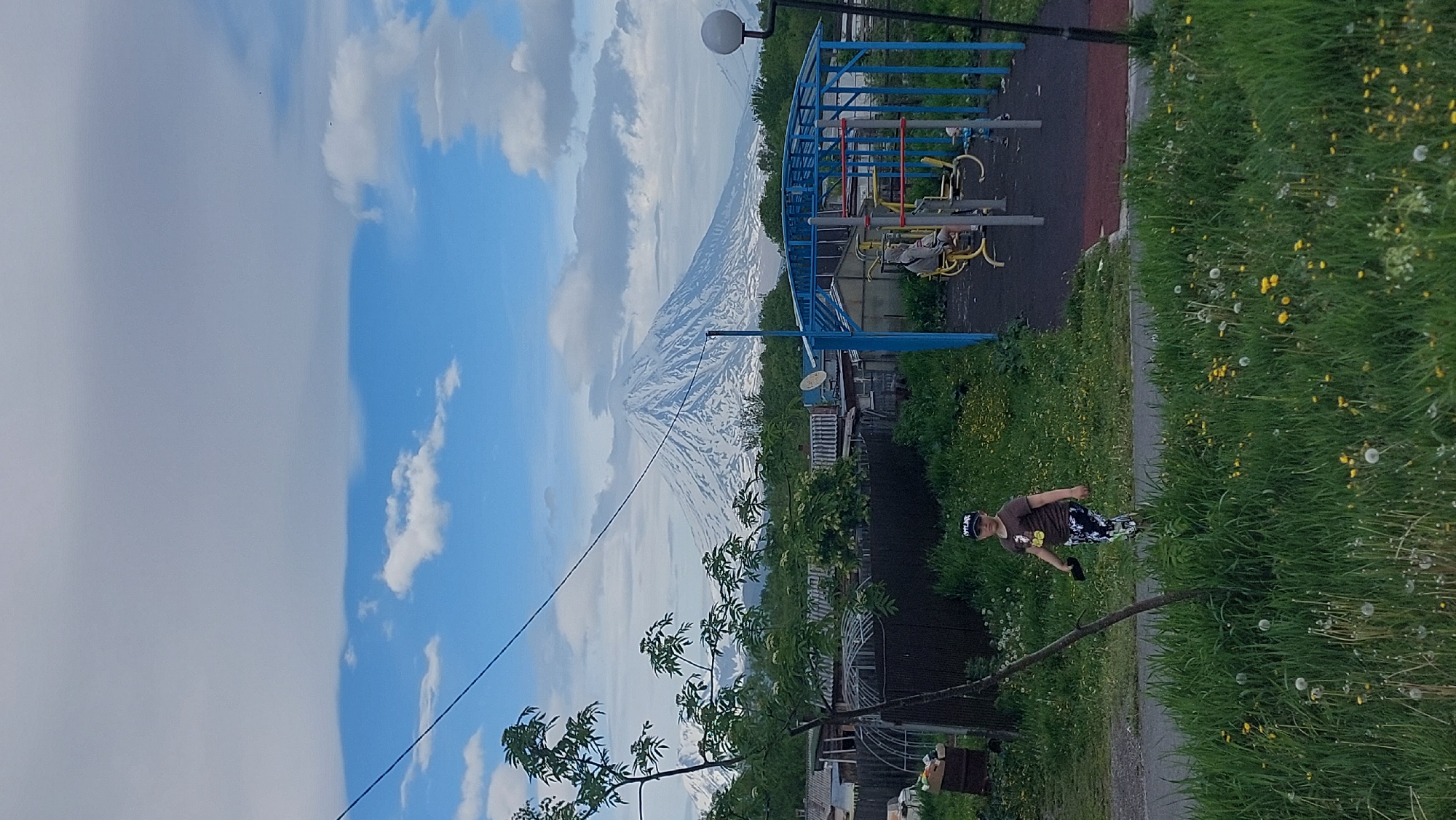 A young boy wearing a dark t-shirt and patterned pants walks across a grassy area in front of a residential neighborhood. He is facing forward and holding a dark object in his right hand. Behind him, in the middle ground, are several low-rise buildings and a blue metal playground structure with exercise equipment. In the background, a large, snow-capped volcano dominates the landscape under a partly cloudy sky. The scene appears to be located in a rural or semi-rural area, possibly in the vicinity of a city like Yelizovo, near Petropavlovsk-Kamchatsky, Russia, given the presence of volcanoes.