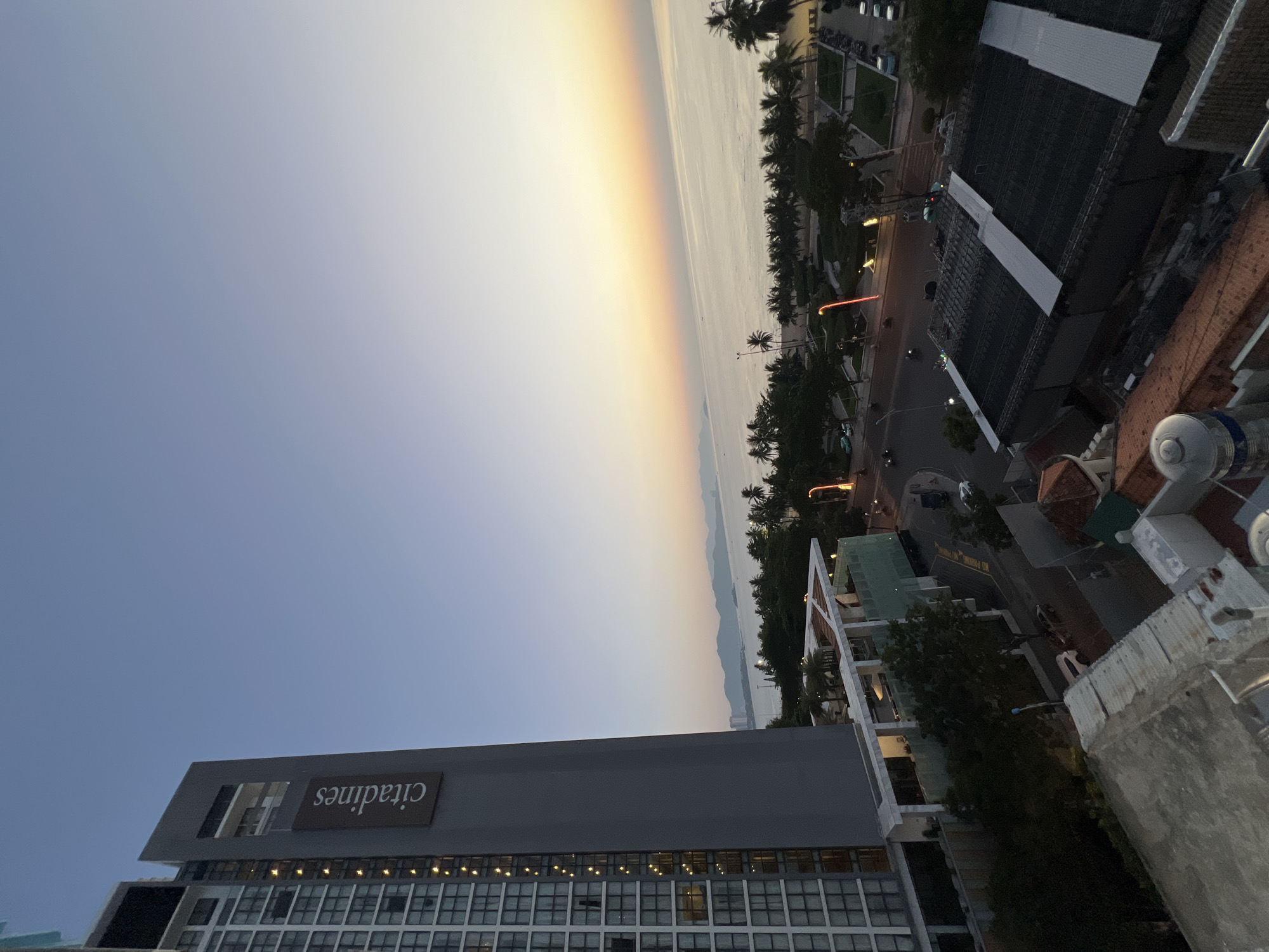 This high-angle exterior shot captures a panoramic view of the coastline in Nha Trang, Vietnam, during a twilight hour, likely sunrise or sunset. Dominating the left side of the frame is a tall, modern hotel building with a prominent "Citadines" logo visible on its facade. The building's glass facade reflects the soft, ambient light, and several windows are illuminated from within.
In the foreground, the rooftops of other buildings are visible, showing a mix of architectural styles, including corrugated metal and tiled roofs. The middle ground reveals a street scene with palm trees lining the beachfront. Vehicles, including cars and motorcycles, are present on the road, indicating active urban life. Lights from street lamps and buildings cast a warm glow.
The background features a serene body of water, likely the South China Sea, with a distant coastline and mountains visible on the horizon. The sky transitions from a pale blue overhead to soft hues of yellow and orange near the horizon, characteristic of dawn or dusk. The overall atmosphere is peaceful, with a sense of urban development meeting natural coastal beauty. There are no distinct people or specific actions readily identifiable in the image beyond the general presence of vehicles and activity.