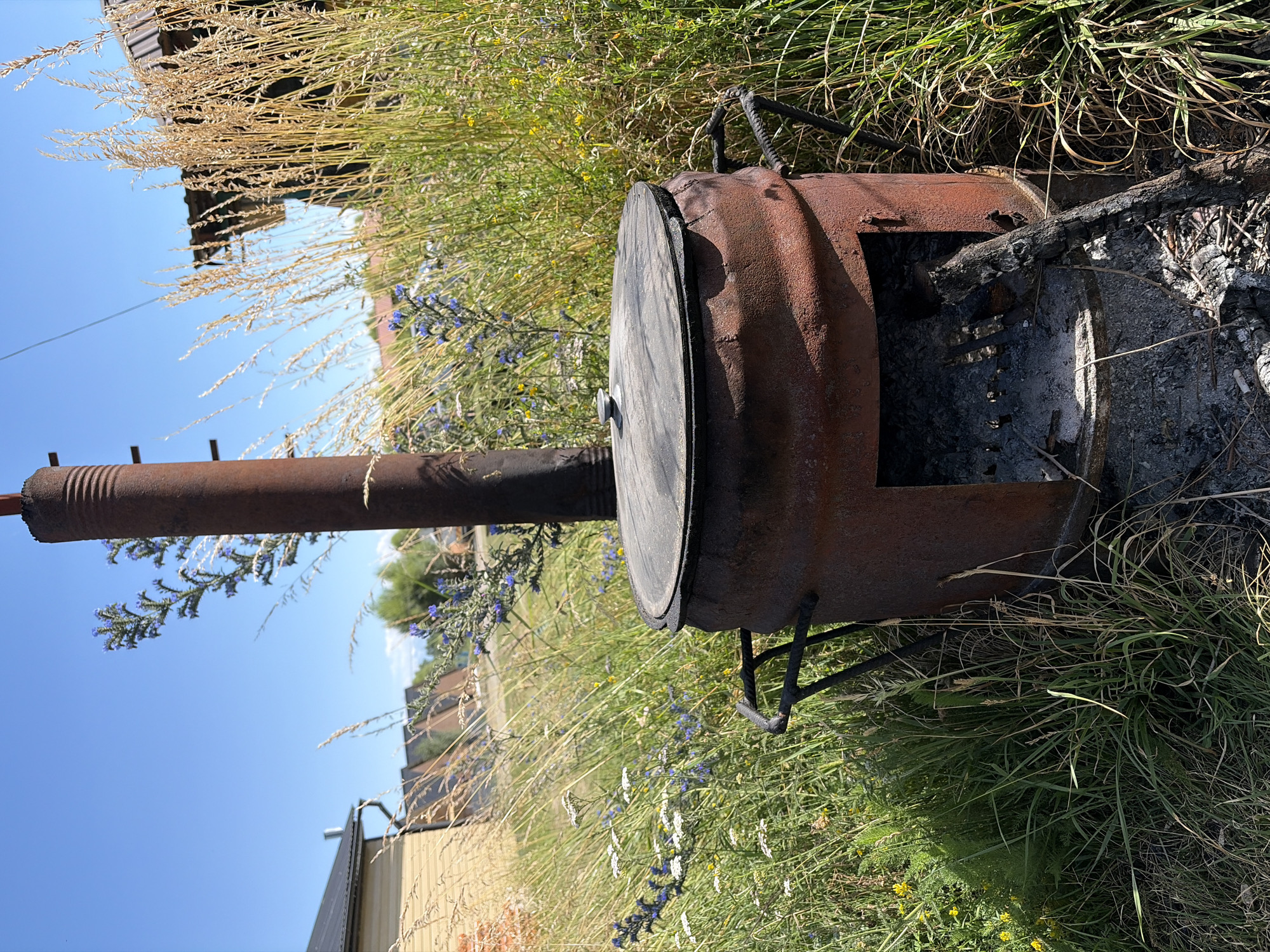 An outdoor shot captures a rusty, cylindrical metal stove with a thick metal lid. The stove has an open firebox at its base, where ashes and burnt wood are visible. A tall, dark metal pipe extends vertically from the top of the stove. The stove is situated amidst tall green grass, wild flowers, and dried stalks of wheat. In the background, wooden houses with dark roofs and a clear blue sky are visible.