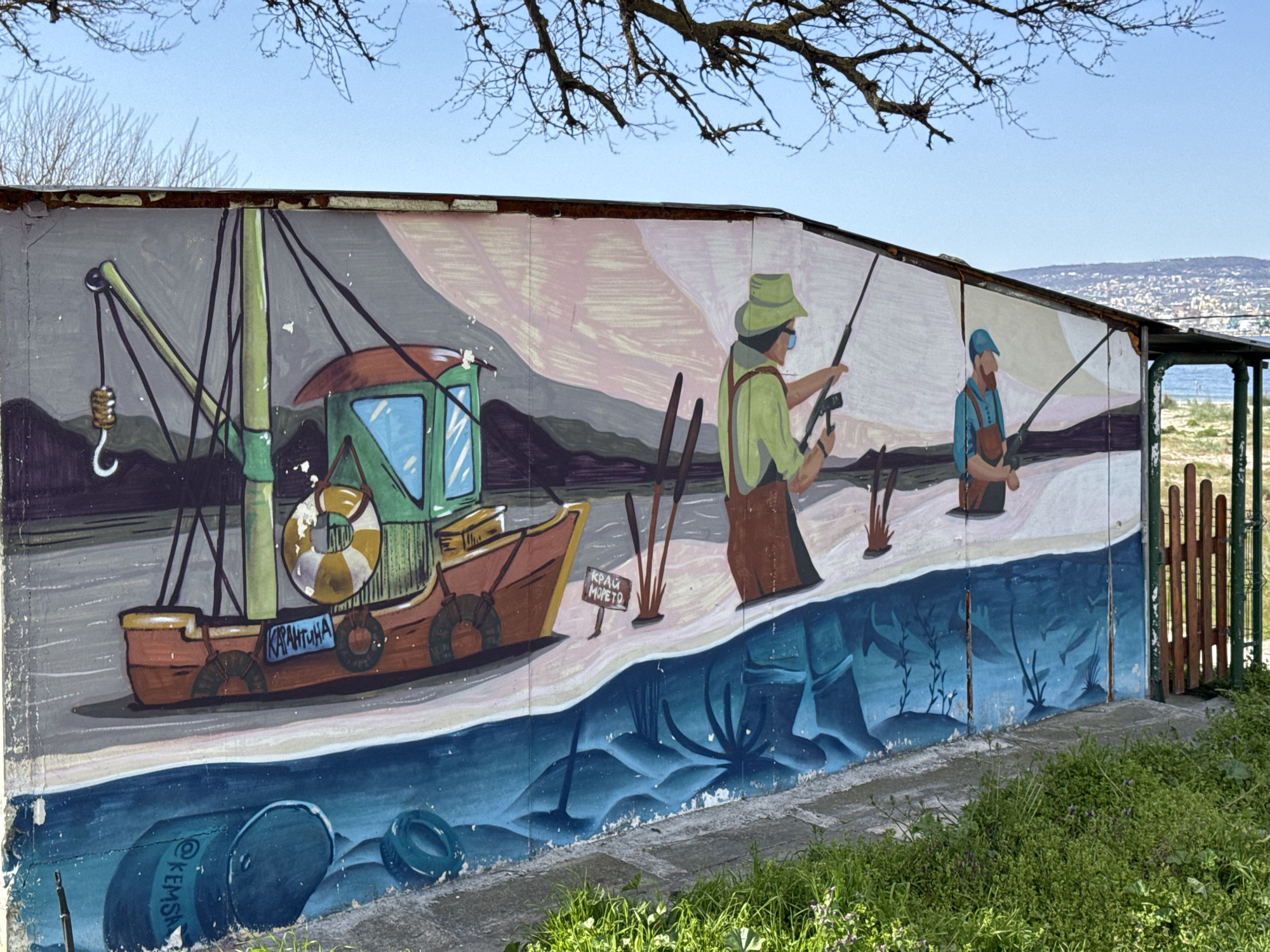 A vibrant mural depicting a fishing theme is painted on a long, low concrete wall, likely an outbuilding or fence, under a clear, bright blue sky. Bare tree branches extend across the top of the frame.
The mural itself is divided into sections. On the left, a cartoonish red and green fishing boat with a large crane/mast is depicted. It features lifebuoys and what appears to be a cabin. Visible text on the boat reads "КАРАНТИНА" (KARANTINA).
To the right of the boat, two men are shown fishing. The first, on the left, wears a light green shirt, brown overalls, and a wide-brimmed green hat, holding a fishing rod. The second man, further right, wears a blue shirt, brown overalls, and a blue cap, also holding a fishing rod. A small signpost between the boat and the first fisherman reads "КРАЙ МОРЕТО" (KRAY MORETO), which translates to "By the Sea" or "End of the Sea." The background behind the figures shows stylized, undulating landforms in muted tones.
The lower part of the mural simulates water, rendered in varying shades of blue, with darker tones representing depth. Submerged objects resembling barrels or tires are visible, one of which has "KEMSΛ" (likely KEMSA) written on it. Silhouettes of fish and underwater plants are also depicted in this watery section.
In the foreground, lush green grass and weeds grow along the base of the wall. On the far right, a rustic wooden gate provides a glimpse beyond the wall to a body of water, possibly the sea, and a distant city or town sprawling across a hillside under the bright sky. The scene suggests a sunny day, likely morning or early afternoon, in a coastal setting in Varna, Bulgaria.