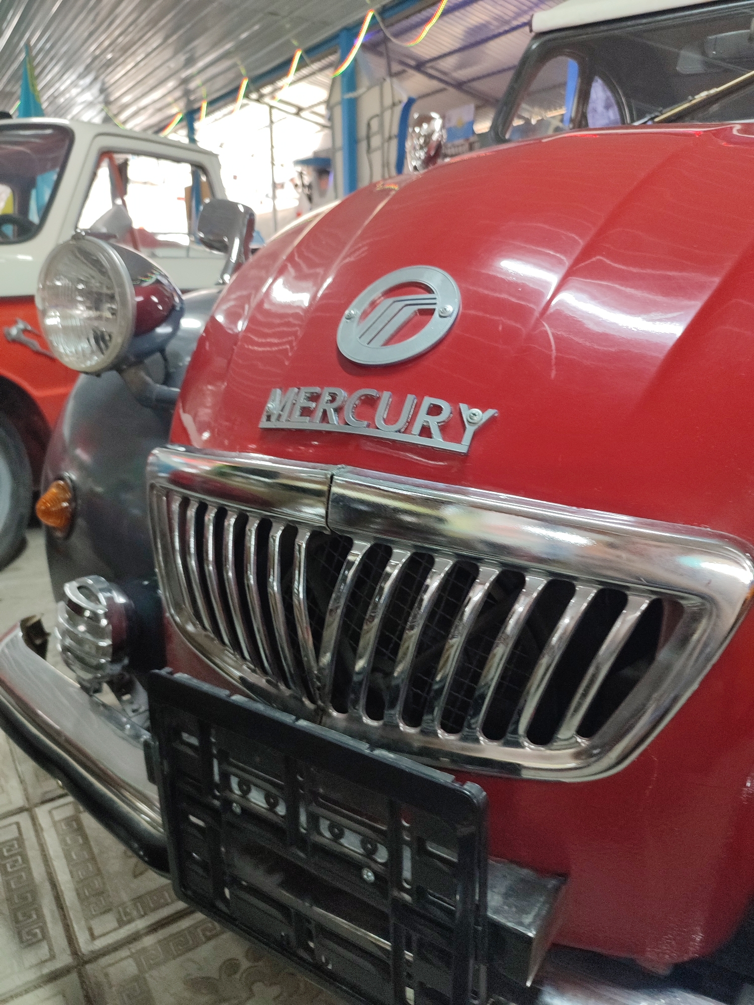 A close-up, low-angle shot captures the front of a vintage red Mercury car. The car's prominent chrome grille, featuring vertical slats, is centrally positioned. Above the grille, the "MERCURY" wordmark is clearly visible in silver lettering, accompanied by the Mercury emblem, a silver winged circle, to its left. A black license plate bracket is attached to the lower front of the car. To the left of the red car, the front of a white and dark grey car is partially visible, including a round headlight. The cars are indoors, likely in a garage or exhibition space, with a corrugated metal ceiling and colorful festive decorations hanging overhead. The floor appears to be tiled with a geometric pattern. This image was taken in the city of Qonaev, Kazakhstan.