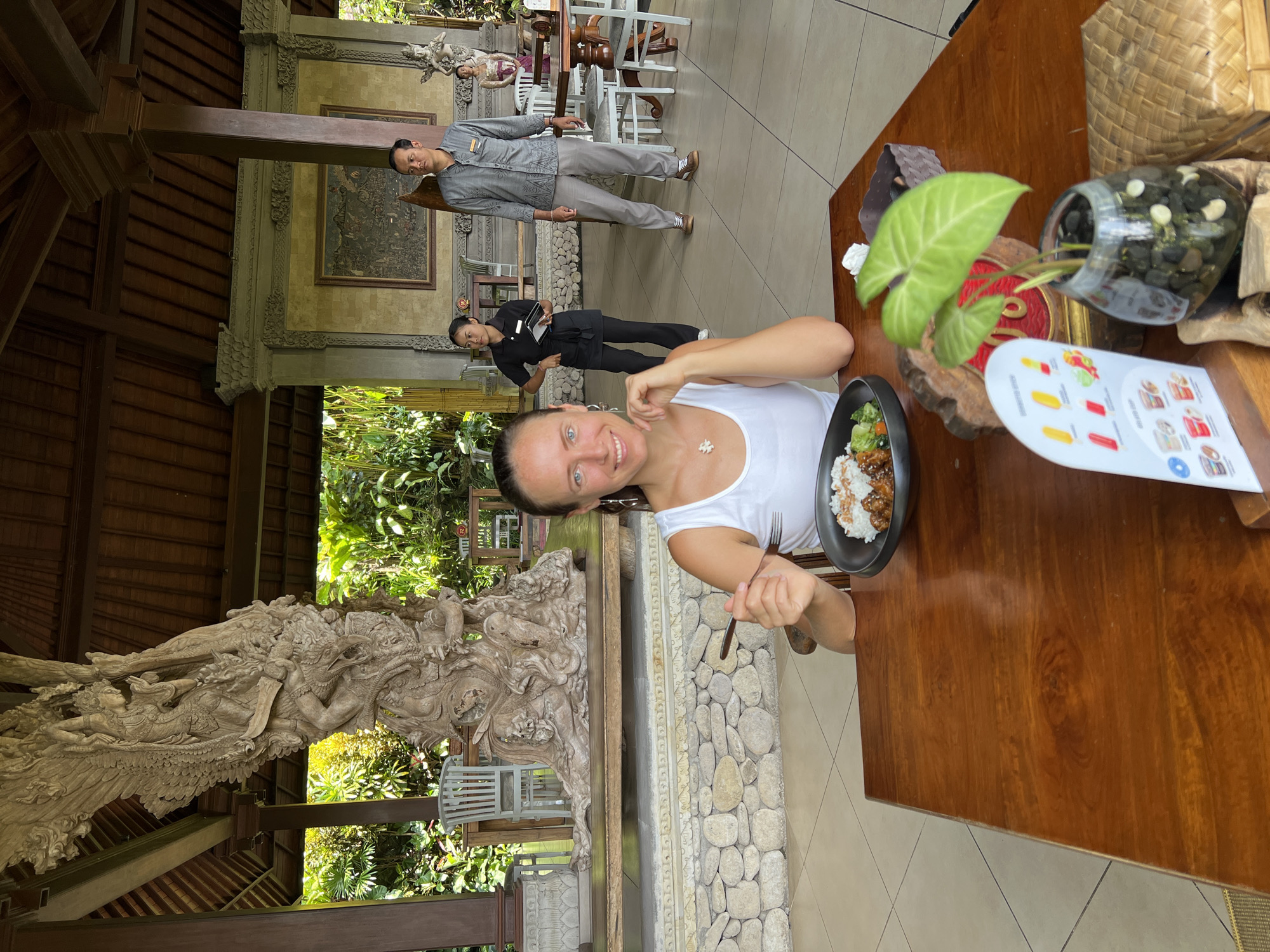 A woman sits at a wooden table in a restaurant in Kedewatan, Indonesia. She is wearing a white tank top and holds a fork. On the table is a plate of food, a plant in a glass vase filled with rocks, a menu, and a woven basket. A waiter and a waitress stand behind her. In the background is a detailed stone carving, lush greenery, and an open-air dining area.