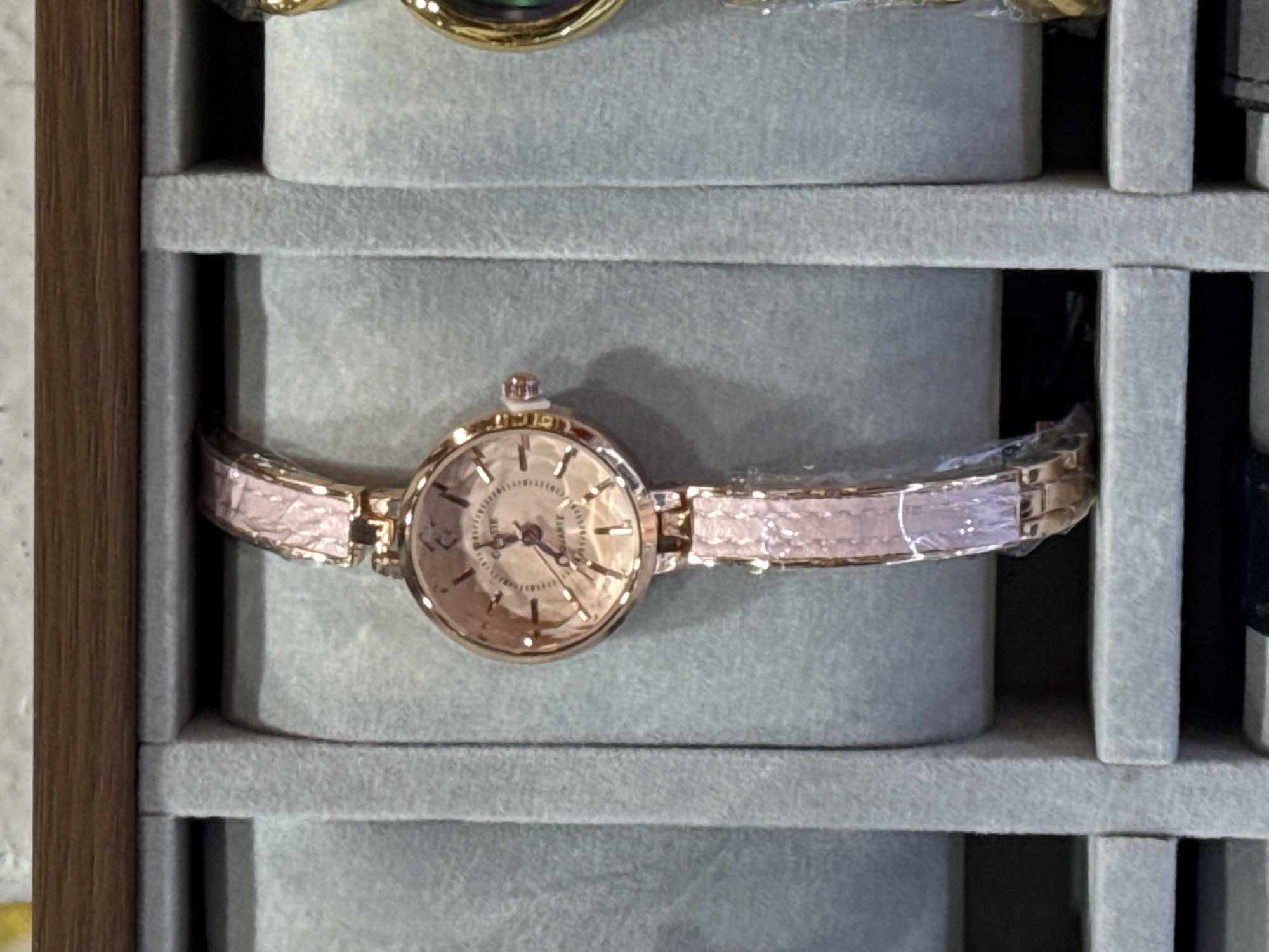 A wristwatch is displayed in a gray compartmentalized tray. The watch is rose gold in color with a round face and a metallic band. The tray appears to be part of a larger display case, based on the presence of other objects and the frame of the case. The location is specified as Tay Nha Trang Ward, Vietnam, but this detail is not discernible from the image alone.