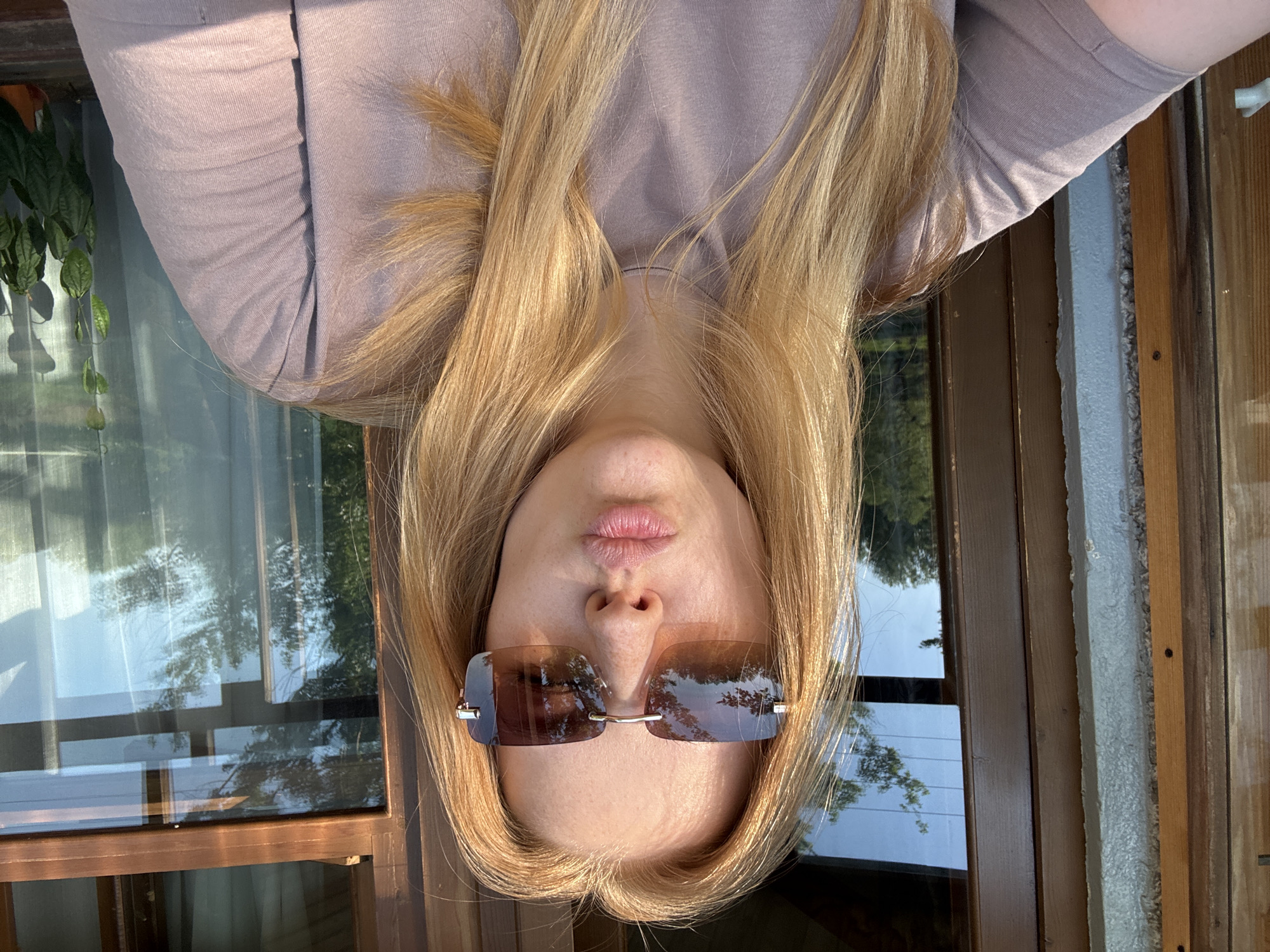 A woman with blonde hair is depicted upside down, wearing brown rectangular sunglasses. The reflection on the sunglasses reveals trees and sky. She is wearing a light brown t-shirt. Behind her is a window and wooden structure. The location is possibly Rusanovo, Russia.