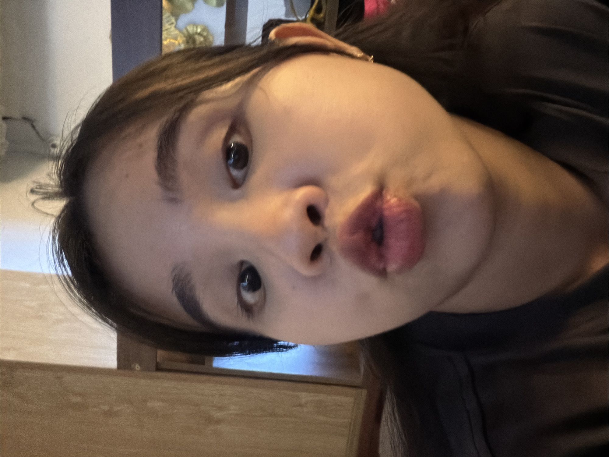 A close-up portrait-style photograph features a young person, likely female, captured from the chest up in a playful pose. The subject has dark hair, dark eyes, and fair skin, and is making a distinct "pouty" or "kissing" face with full, pursed lips, while looking directly at the camera. A small earring can be seen on their right ear. They are wearing a simple, dark-colored top, possibly black or dark gray.
The background elements suggest an indoor setting, with a blurred light-colored wall or surface behind the person's head. Hints of decorative, possibly gold-toned, abstract elements are visible further back on the right side of the frame. Below the chin, a wooden surface, resembling a table or cabinet, is present. The lighting is soft and frontal, illuminating the subject's face with a warm glow. The overall mood of the image is expressive and lighthearted. No visible text is present. This scene is set in Đà Nẵng, Vietnam.