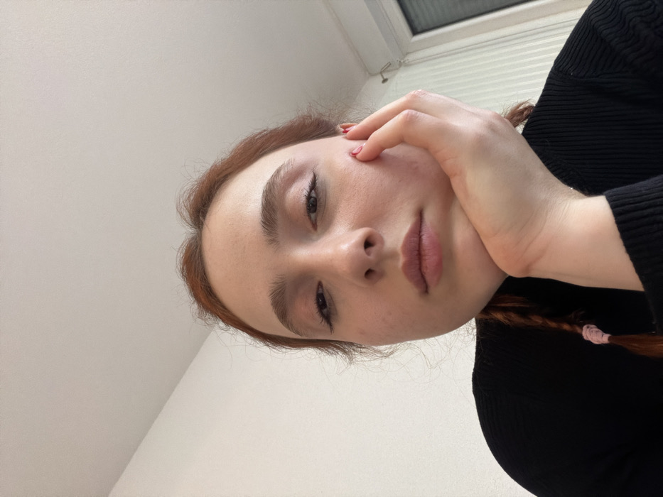 A young adult female with reddish-brown hair is depicted in a close-up, low-angle indoor shot, positioned in Eschau, France. She faces the camera directly, her head tilted slightly, with a neutral expression. Her right hand supports her chin and lower jaw; her fingernails exhibit red polish. She wears a black, ribbed long-sleeved top. A braid secured by a pink elastic tie is visible on her left side. The background consists of a plain, light-colored wall and ceiling, with the upper right portion of a window frame and vertical blinds partially visible on the right. A white bracket or fixture is mounted on the wall near the window.