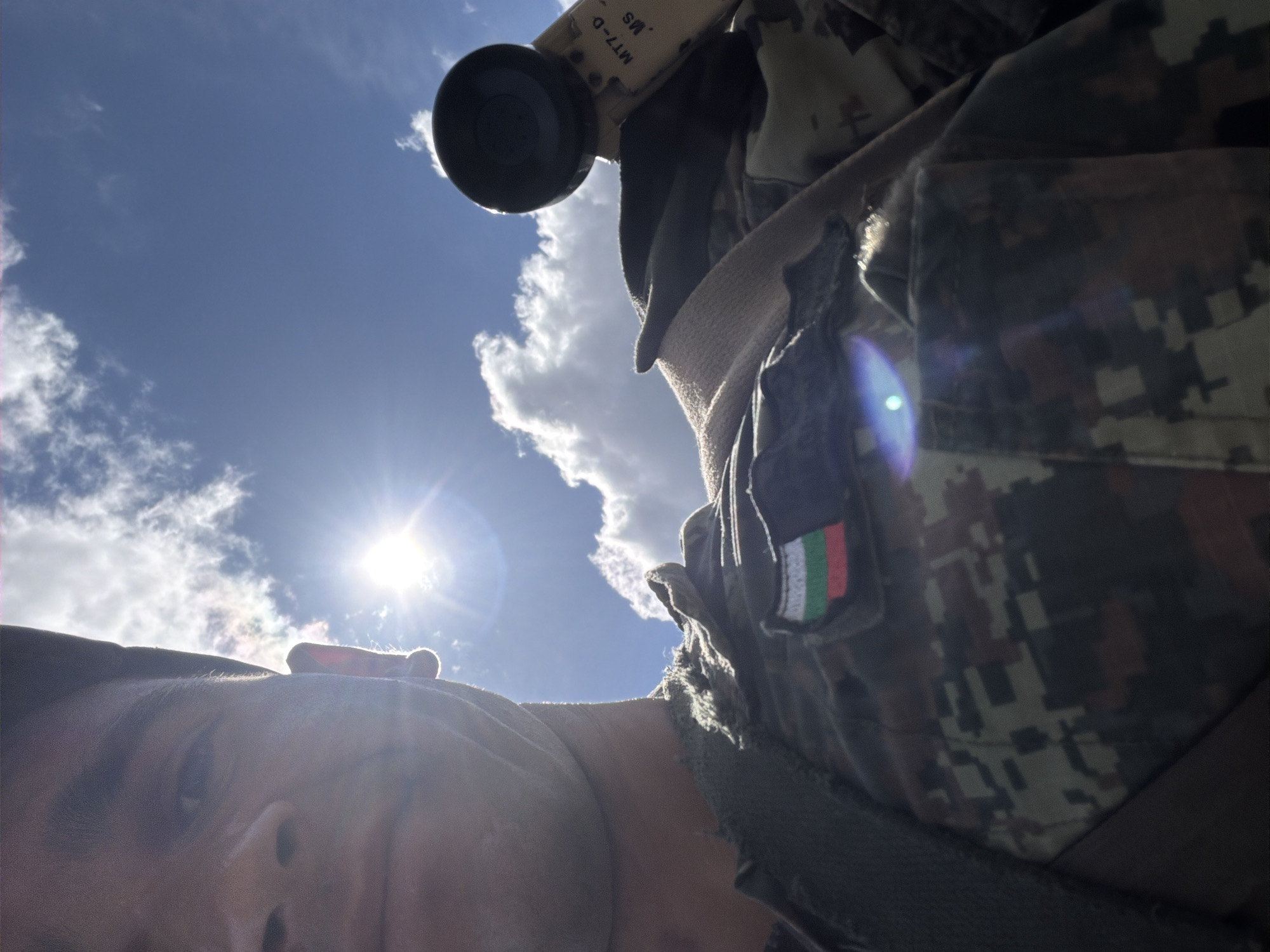 The image is a low-angle, close-up selfie taken outdoors on a sunny day with clouds. The subject, a person wearing a camouflage uniform with a Bulgarian flag patch on the left chest, is partially visible in the foreground, looking upwards. Part of their face, including an ear and cheek, is visible on the left. The person is holding an object with a black circular end and some text, possibly part of a communication device or a weapon sight, on the right side of the frame. The background consists of a bright blue sky with scattered white clouds and the sun shining intensely, creating lens flare. The overall setting appears to be an outdoor environment, possibly a military training area or a field in the vicinity of Beliakovets, Bulgaria, given the presence of the Bulgarian flag.