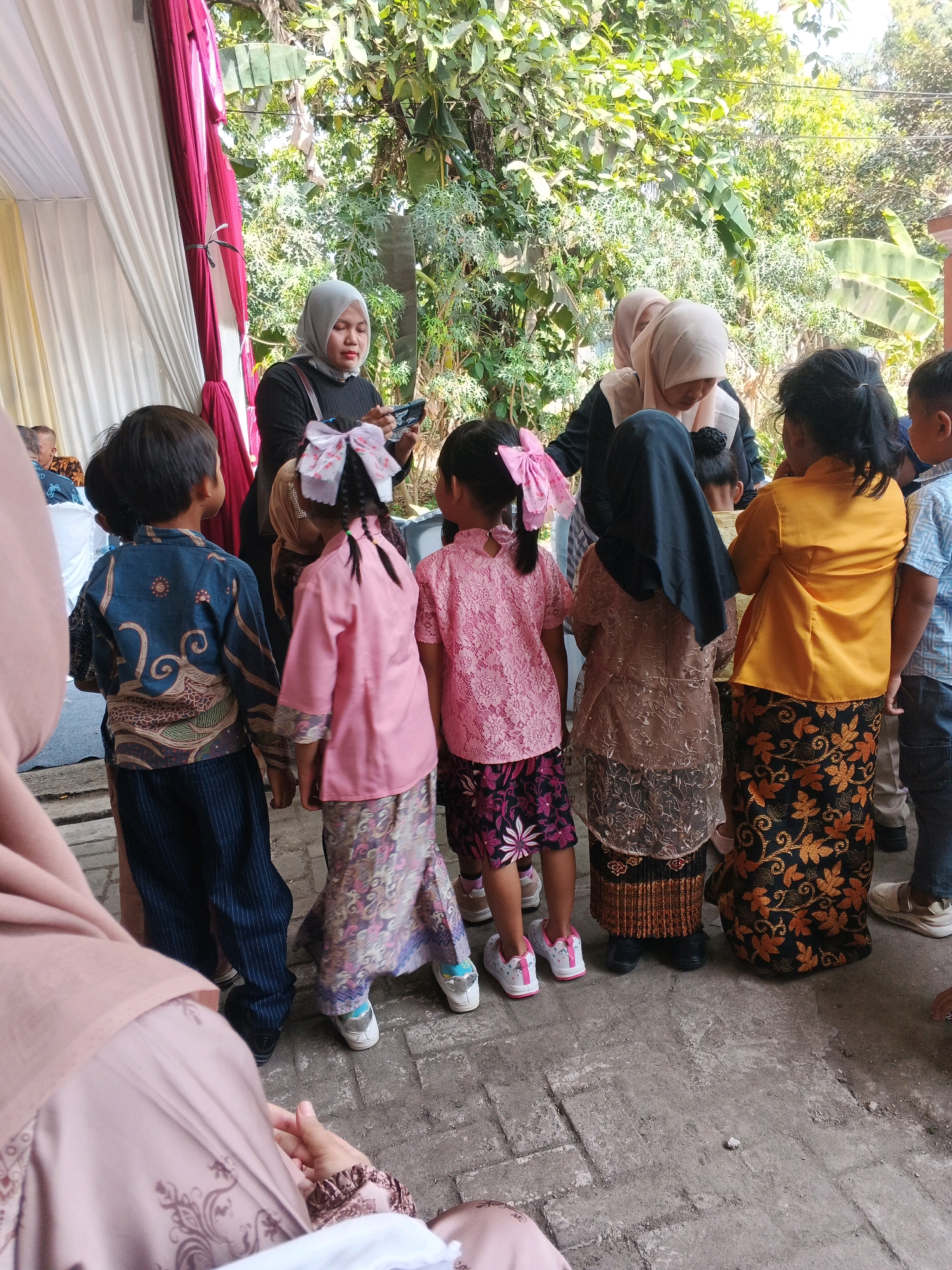 In Kadu, Indonesia, a group of children and adults are gathered outdoors, possibly for an event, under dappled sunlight filtering through lush green trees. Several children, all wearing traditional Indonesian clothing, stand facing away from the camera. One child in a pink top and patterned skirt has a large pink bow in her braided hair. Another, in a blue patterned top and dark pants, stands to the left. An adult woman, wearing a grey hijab and black top, holds a phone and appears to be interacting with the children. Another adult, also in a hijab, is partially visible behind the children. The ground is paved with rough concrete tiles. A white and maroon draped tent or fabric structure is visible on the left side of the frame.