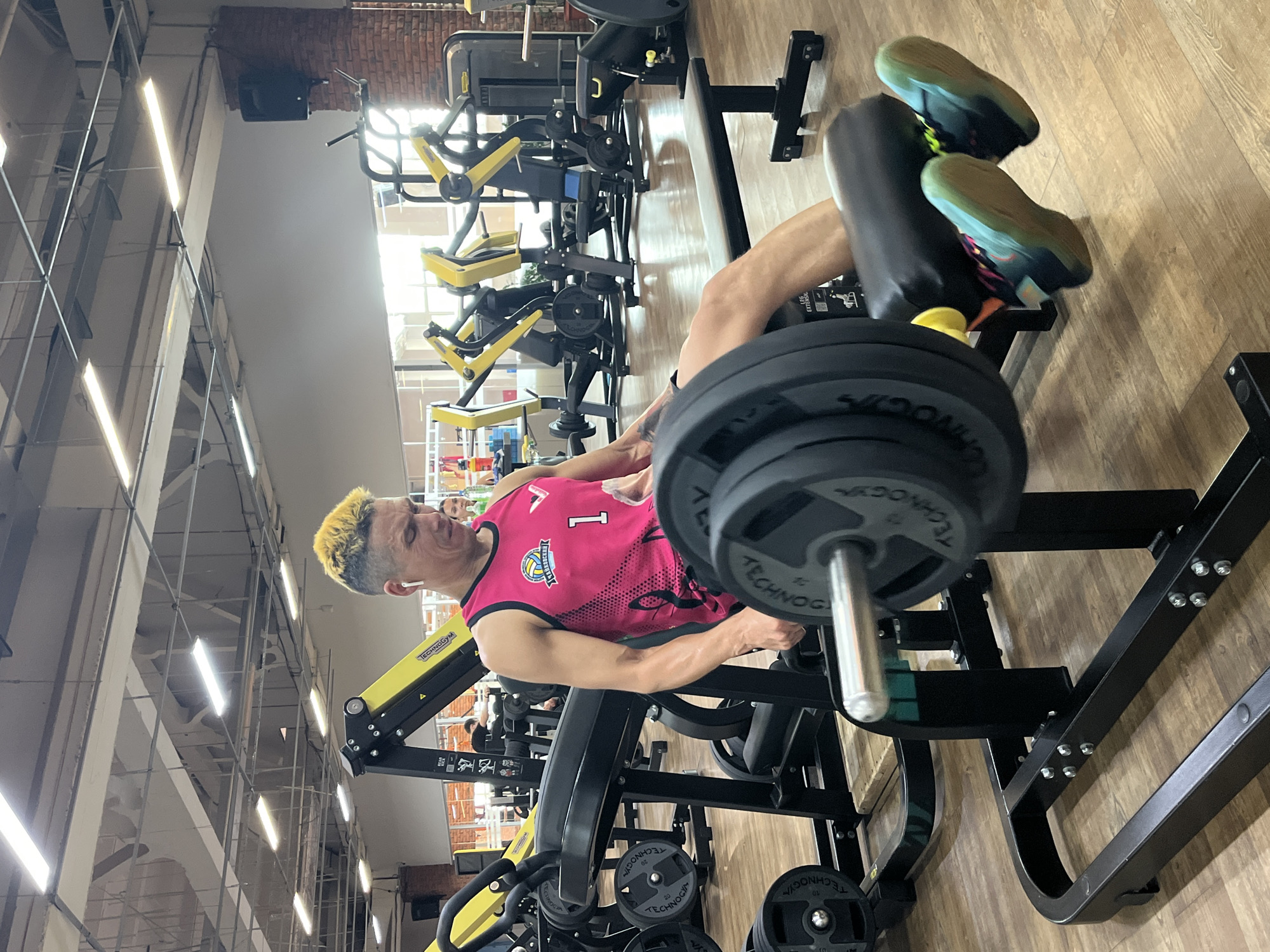 This image depicts a man performing a leg extension exercise on a weight machine in a gym. The man has brightly colored yellow and black hair and is wearing a pink tank top with a logo that includes a volleyball and the number "1". He is also wearing wireless earbuds. The leg extension machine is black and yellow, with weight plates labeled "Technogym". In the background, other gym equipment, including more weight machines and possibly treadmills, is visible, along with windows that suggest daylight. The flooring appears to be wood. The gym is located in Chaikovsky, Russia, indicated by the logo on the man's shirt, which features Cyrillic text and likely represents a local sports team or club.