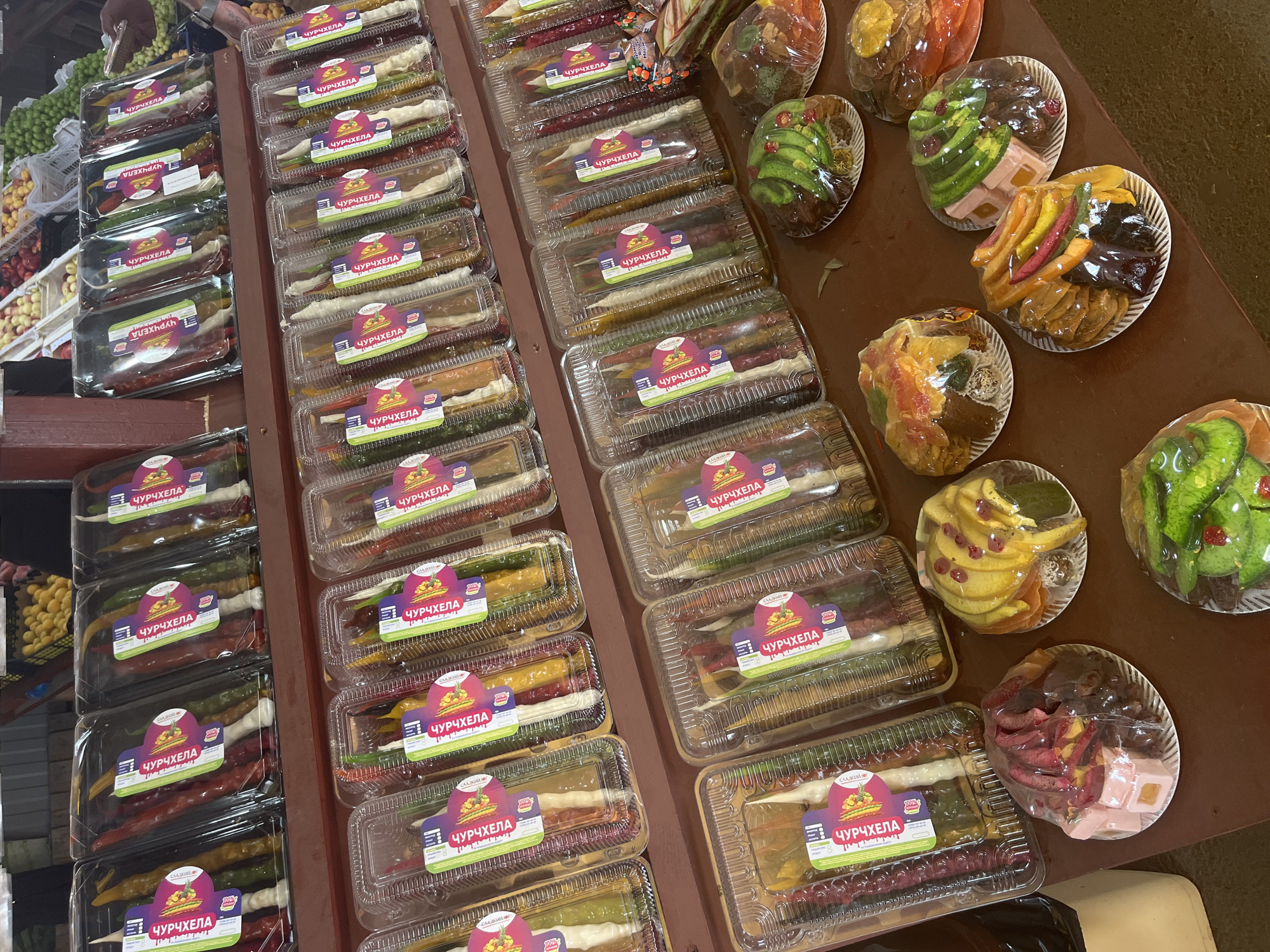 The image shows a stall at an outdoor market in Plastunovskaya, Russia, displaying a variety of packaged and unpackaged dried fruit candies. On shelves in the background, rows of long, thin, multi-colored candies, identified by labels in Cyrillic as "ЧУРЧХЕЛА," are neatly arranged in clear plastic packaging. These candies appear to be made of nuts coated in a fruit-based jelly. In the foreground, on a table, are several plates of colorful dried fruit and candies, also wrapped in plastic. These plates display a variety of shapes and colors, including what appear to be sliced bananas, figs, apples, and possibly other fruits, some arranged in decorative patterns. The overall impression is of a vendor selling traditional confectionery. No people are visible in the frame. The environment appears to be an open-air market with wooden stall structures.