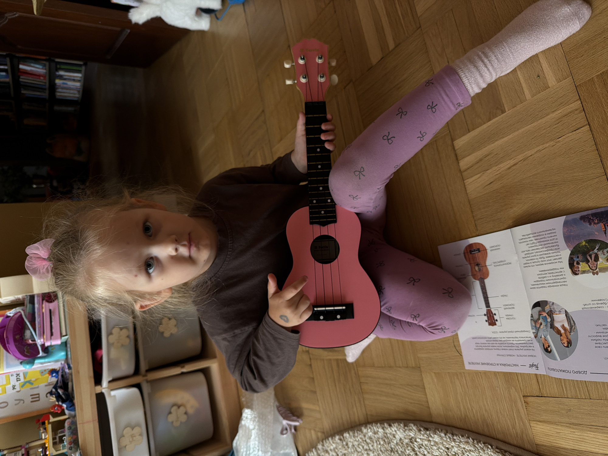 A young girl with blonde hair tied with a pink bow is sitting on a light brown wooden floor. She is wearing a dark brown long-sleeved shirt and purple leggings with a bow pattern. In her hands, she holds a pink ukulele, which is positioned horizontally. Her right hand is on the neck of the ukulele, and her left hand is near the strings. A booklet is laid open on the floor in front of her, displaying images of ukuleles and people. The environment appears to be a room with shelves filled with toys and books in the background. The lighting suggests an indoor setting. The image was taken in Khimki, Russia.