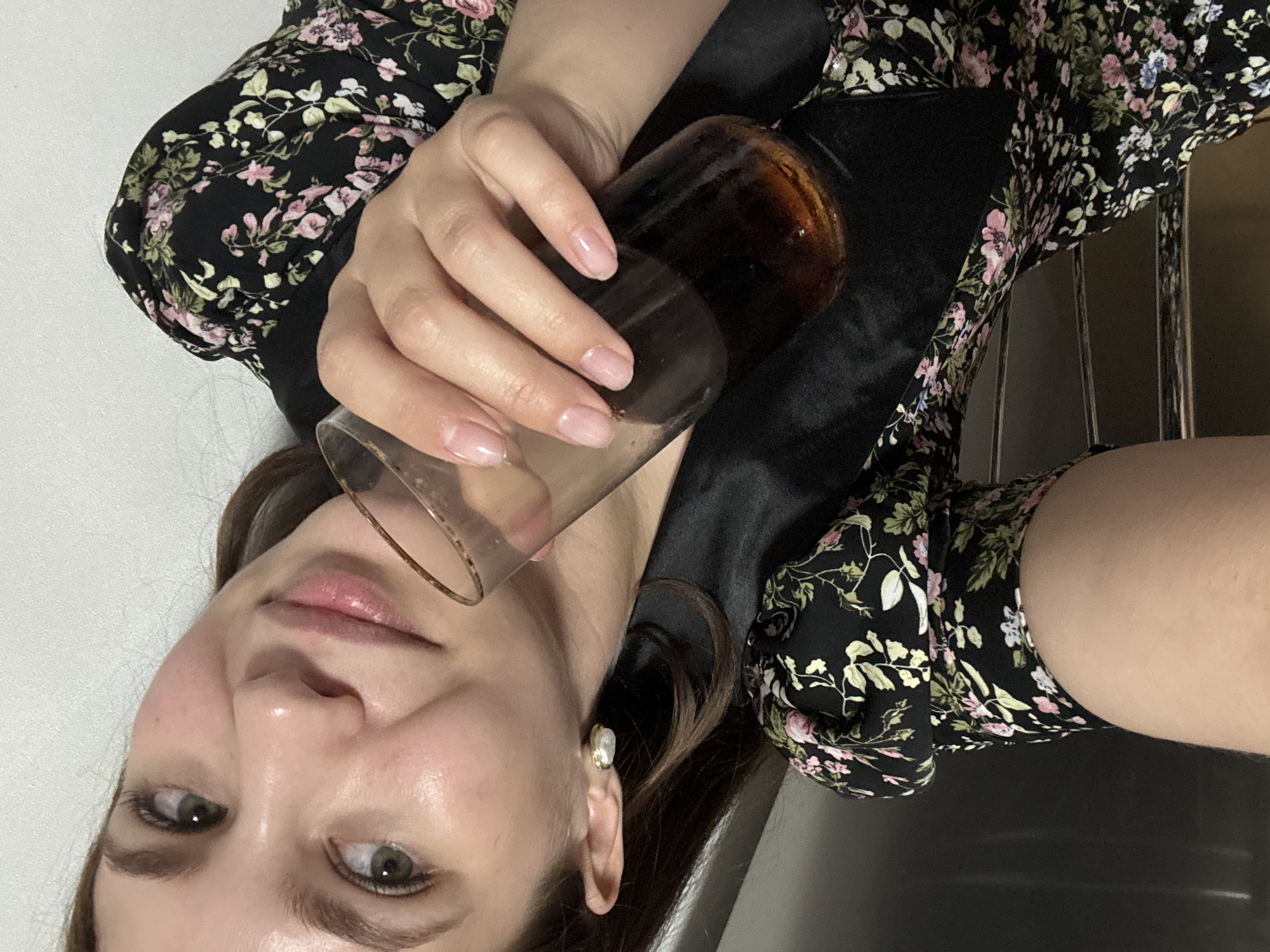 The image is a close-up, selfie-style shot of a young woman lying down. She is wearing a black dress with a floral pattern of pink and white flowers and green leaves. A portion of her left arm is visible, and she is holding a glass of dark liquid, likely soda, in her right hand. The glass has a condensation ring on its rim. The woman has light skin, brown hair, and light green eyes. She is wearing a pearl earring on her left ear. Her fingernails are painted a light pink color. The background appears to be a plain white wall. The image appears to be taken indoors. No specific landmarks or contextual clues are visible to indicate a location in Chaikovsky, Russia.