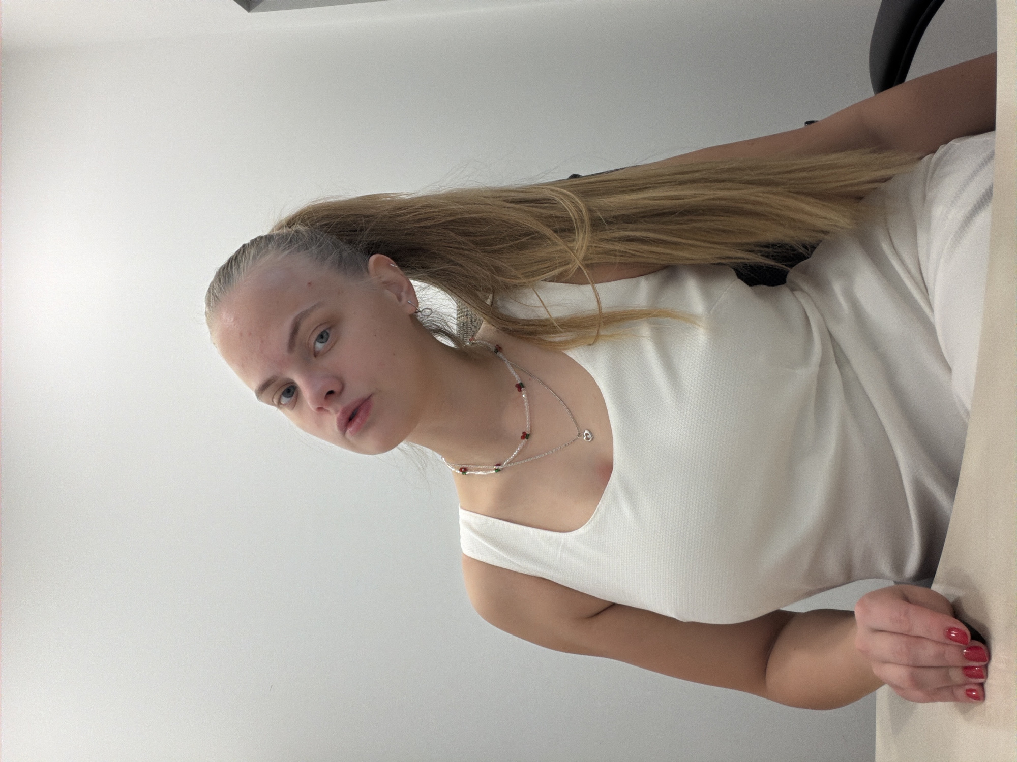 A young woman with blonde hair pulled back in a ponytail is sitting at a table in what appears to be an office or home setting. She is wearing a white sleeveless top and a delicate necklace. Her nails are painted red. The background is a plain white wall. There's no indication that the image was taken in Izhevsk, Russia.