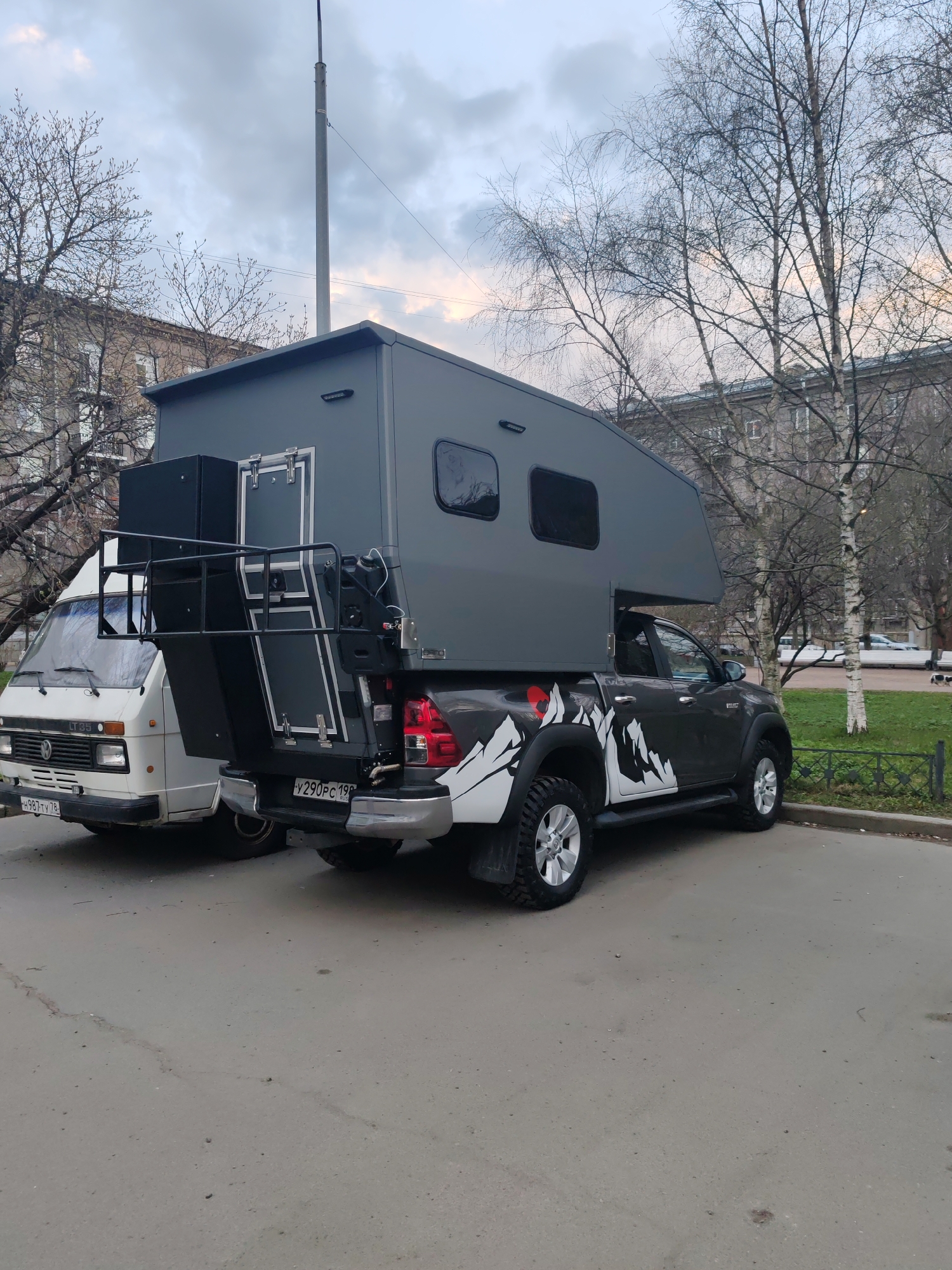 The image presents two customized vehicles parked on an asphalt surface in an urban setting, likely Saint Petersburg, Russia, given the visible license plate region codes (198 and 78). The sky is overcast, and trees with bare branches suggest either early spring or late autumn.
The main subject is a dark gray pickup truck, identifiable as a Toyota Hilux from subtle badging, equipped with a large, rectangular, gray camper unit mounted on its bed. The truck features distinctive white, stylized mountain graphics along its rear side panels, including a small red circular emblem on the driver's side. The camper unit has small, rectangular windows and a complex rear section, incorporating a black framed platform with railings and a door, suggesting an access point. The truck's rear license plate reads "Y290PC 198 RUS".
Parked directly behind the customized truck is an older model white van, a Volkswagen LT 35, with its model number visible on the front grille. Its front license plate reads "H987TY 78".
In the background, bare-branched trees, some with light-colored bark typical of birch, stand in front of multi-story buildings. A tall, gray utility pole rises into the sky. The vehicles are stationary, and there are no people visible, suggesting a quiet moment on a street or parking lot. A low, dark fence and a patch of grass are visible to the right. The diffused light indicates daytime under cloudy conditions.