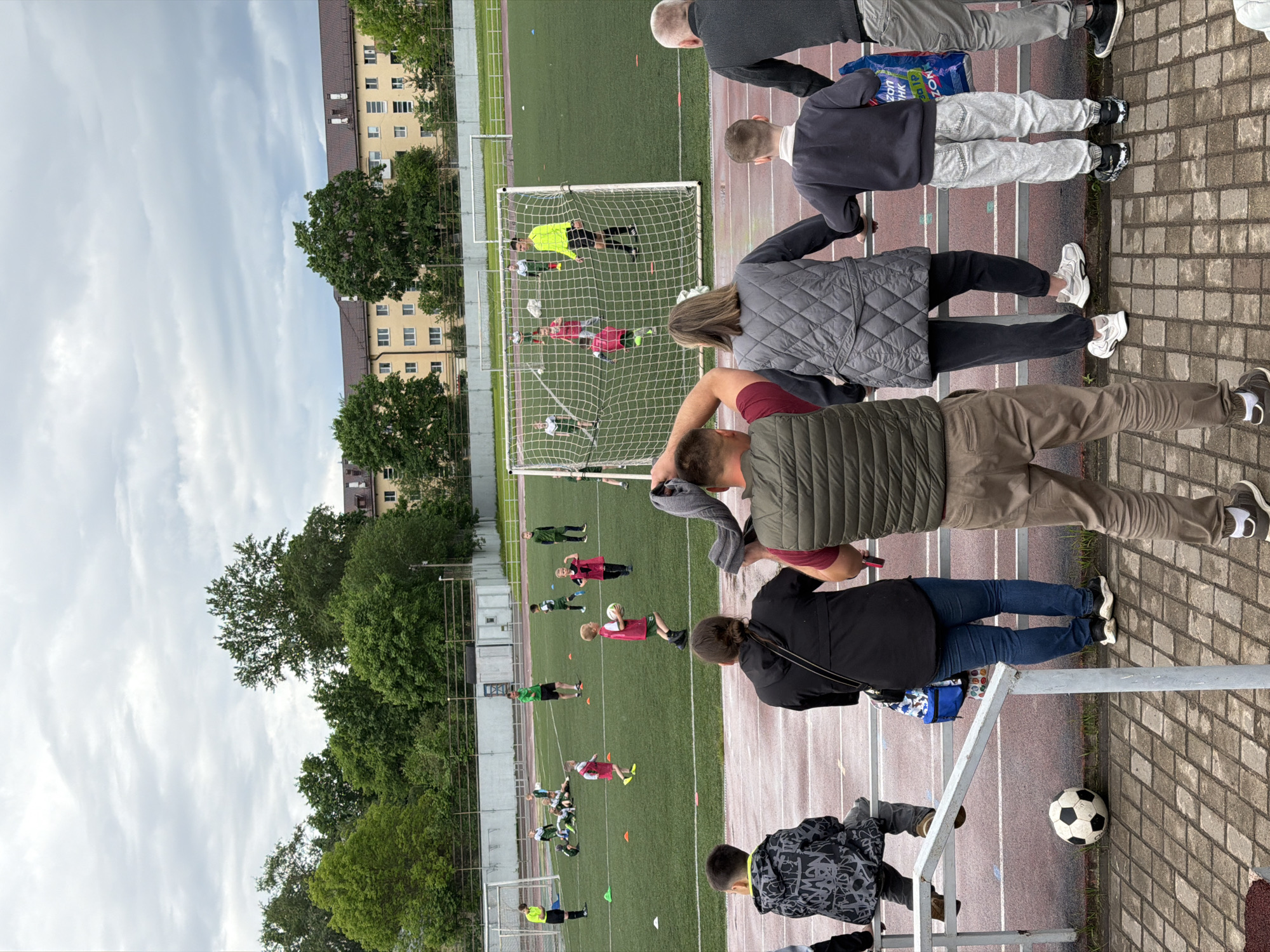 The image shows a soccer game being watched by several spectators. The game is taking place on a green soccer field with white lines and orange cones. The soccer goal has a net. Players in green and red uniforms can be seen on the field. A building with multiple windows and trees surrounds the soccer field. A group of spectators watches the game from behind a metal railing. The spectators are wearing casual clothing and holding their phones. A soccer ball rests on the ground. The image was likely taken at the MBU "Center for Physical Culture and Sports" at the street address of Bolshaya Moscowskaya Street, 108, Veliky Novgorod, Novgorod Oblast, Russia, 173003.