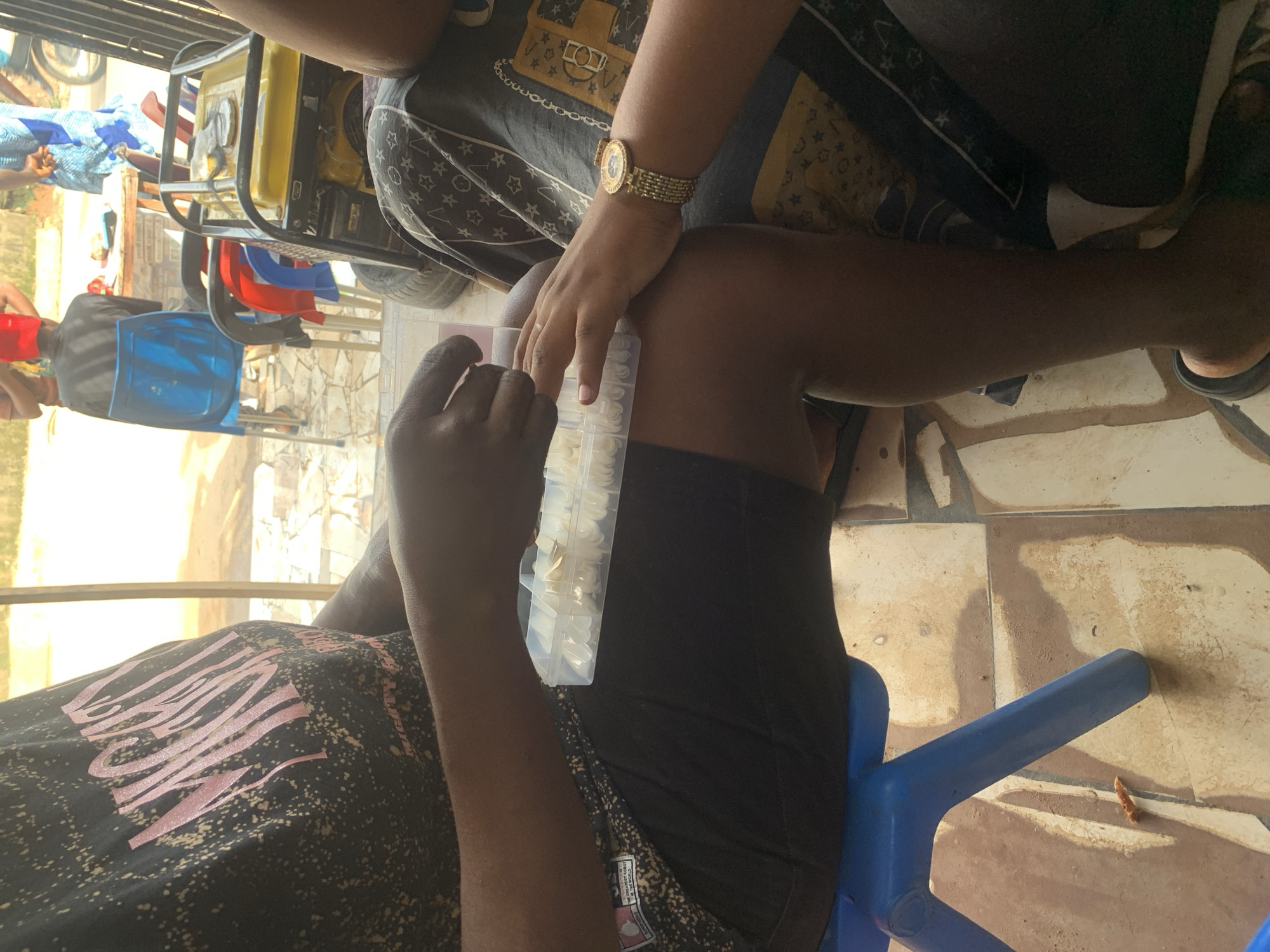 Two people are engaged in a manicure procedure. One person holds a box of artificial nails, while the other prepares to attach them. The scene takes place outdoors, possibly in a residential area in Abuja, Nigeria. Visible elements include: two people, a manicure process involving artificial nails, clothing, chairs, a tiled floor, and a generator. A building and other individuals are in the background.