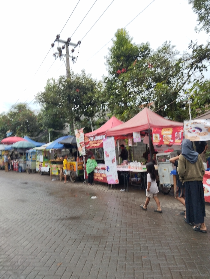 The scene depicts a street food market in Banjaran, Indonesia, located at coordinates -7.047 latitude and 107.58 longitude. The market is comprised of several stalls, each with a red canopy. There are various individuals interacting with the stalls, including vendors, shoppers, and onlookers. The vendors are preparing and displaying food items for sale. The shoppers are browsing the food selection and interacting with the vendors. Some individuals are seated at tables while others are standing or walking. There are various food items displayed for sale, including snacks, drinks, and cooked meals. The market is located on a paved street. The weather is overcast. The street is wet, suggesting that it may have recently rained. The scene is bustling with activity. Trees can be seen in the background, providing shade for the market. Several electrical wires are visible overhead.