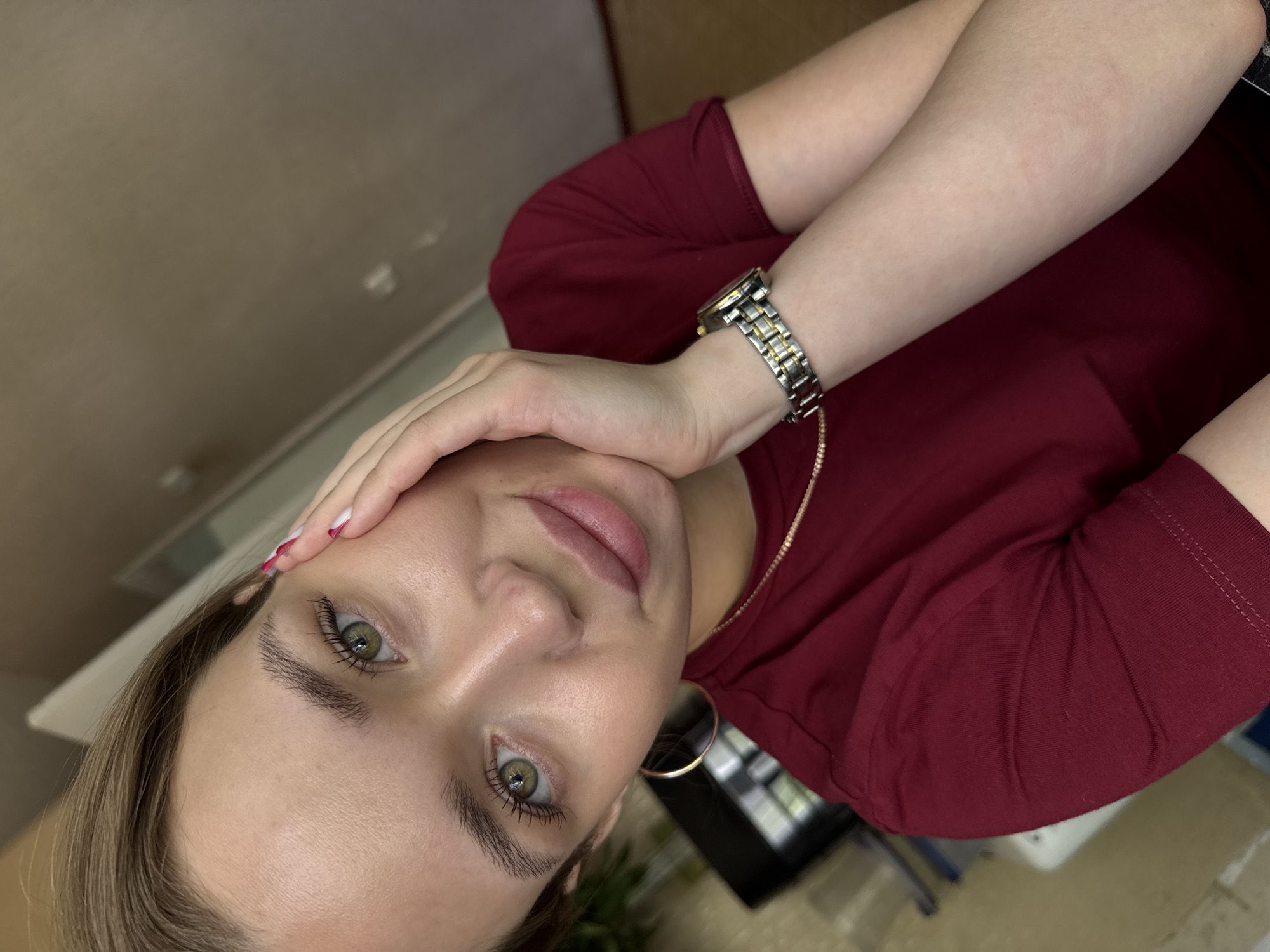 The media file is a close-up, tilted selfie of a young woman indoors. She is wearing a maroon t-shirt and a silver and gold watch on her left wrist. Her right hand is touching her forehead, and her left arm is bent, resting on her chest. She has green eyes and light brown hair. Her lips are slightly parted, and her expression appears neutral or contemplative. The background is out of focus, showing a beige wall and hints of furniture. The lighting suggests it is daytime. There is no visible text in the image. The location context is Chaikovsky, Russia.