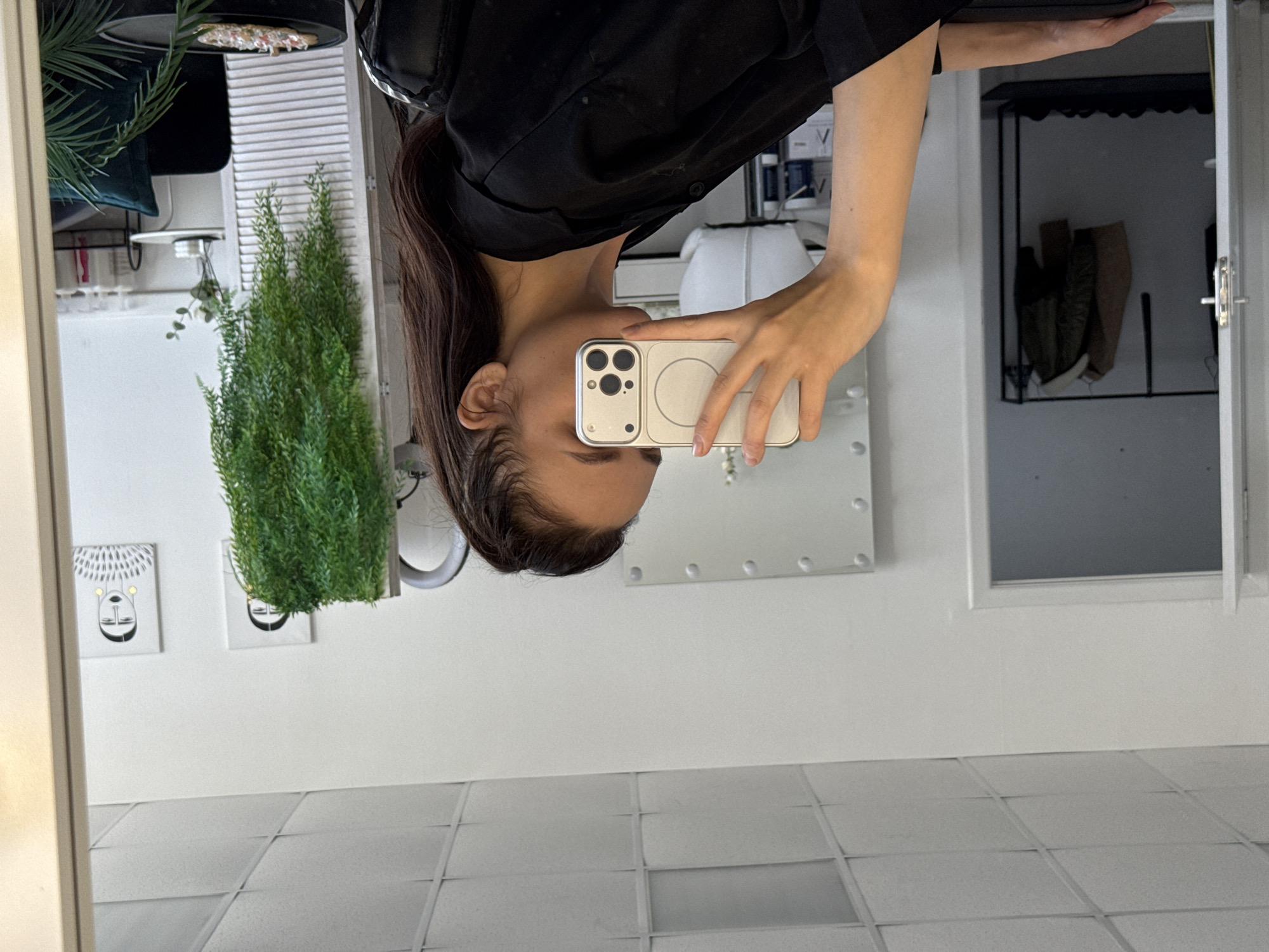 A woman is reflected in a mirror in a bathroom. She is holding a white smartphone in her left hand and taking a selfie. She is wearing a black shirt. In the background, there are decorative plants, pictures, a white storage unit, and tiled flooring. A coat rack can be seen inside an open cabinet. The location is Chaikovsky, Russia.