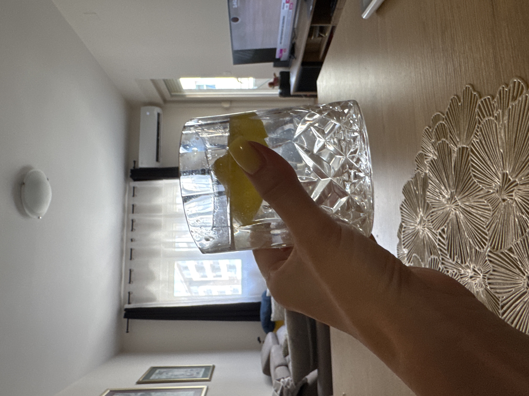 A person's hand, with yellow-painted fingernails, holds a glass of clear liquid with ice and lemon slices. The glass is placed on a wooden table with a decorative placemat. In the background, a living room is visible with a sofa, curtains, a window, an air conditioning unit, and a television. The scene appears to be indoors, likely in an apartment.