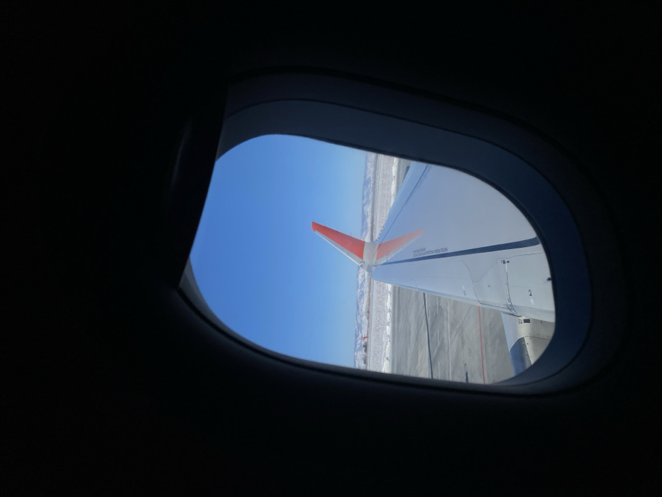 Through an airplane window, a view of the clear blue sky above the plane's wing is visible. The wing features a striking orange winglet. Below, a ground surface, possibly an airport tarmac, appears alongside a snowy landscape. The interior of the plane is dark, contrasting with the bright daylight outside the window.