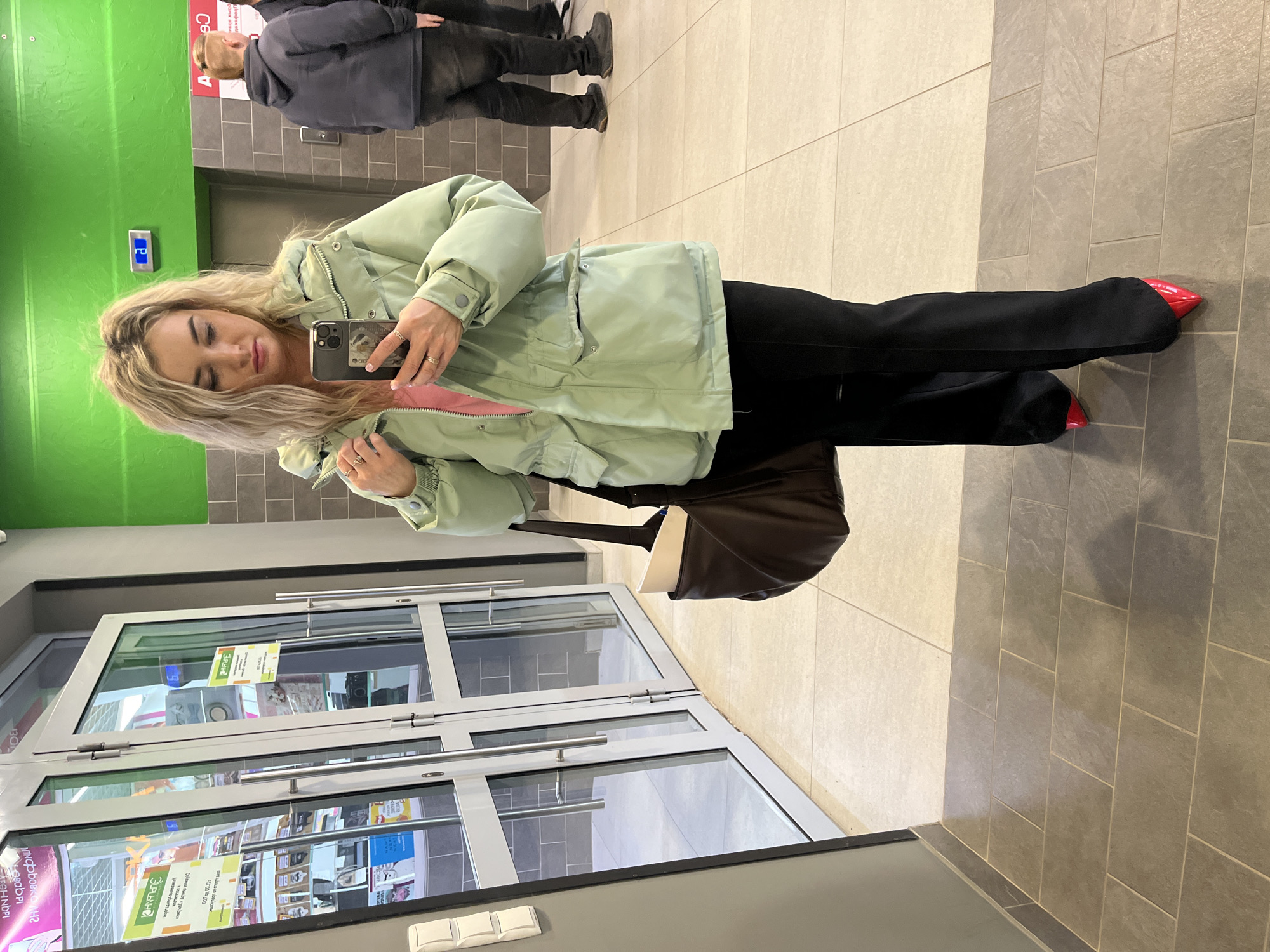 A woman is taking a selfie near the entrance of a store, which is likely in Izhevsk, Russia, based on the Cyrillic script visible on the signage. She's wearing a light green jacket with a pink lining, black pants, and red pointed-toe shoes. She's holding a dark-colored smartphone and has blond hair. Behind her, there's a man in a dark coat and pants, partially visible near an elevator. The background includes a green wall, elevator doors, and tiled flooring. The store entrance has glass doors with silver frames.