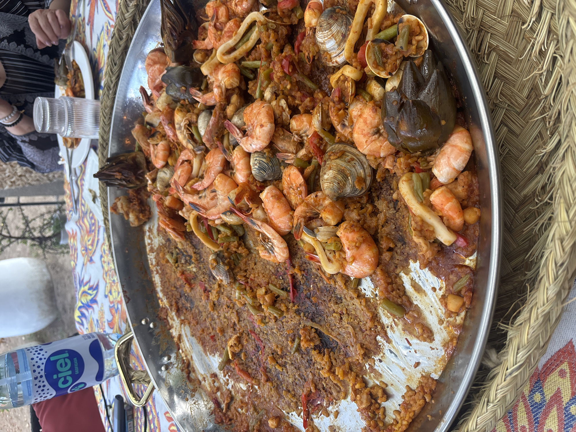 The image shows a large, circular paella pan nearly depleted of its contents, likely a seafood paella containing shrimp, clams, calamari, artichoke hearts, and rice, as evidenced by the remaining portions. The pan sits on a colorful tablecloth. A bottle of "ciel" purified water is also visible. The location of this photograph might be Puebla, Mexico, given the brand of water, typical cuisine, and textiles. A woman's arm is seen at the left of the frame. Another place setting with a glass and fork is also at the left side.