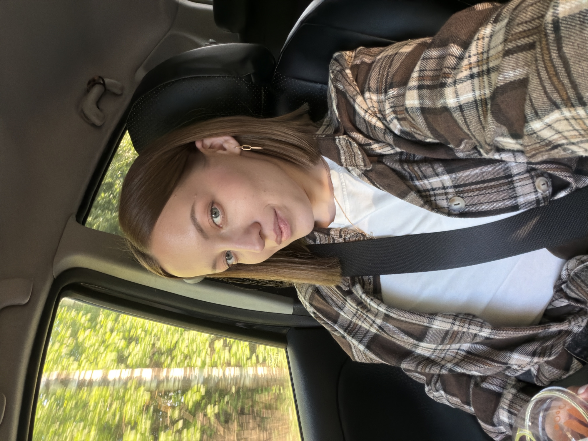 A young woman is sitting in a car. She has short brown hair, fair skin, and is wearing a seatbelt, a white shirt, and a brown and white plaid jacket. Outside the car window, trees and greenery are visible, suggesting a rural or suburban environment. Based on the specified location, the surroundings could be a drive through or near Maksimkovo, Russia. The car's interior is visible, including a black leather seat.