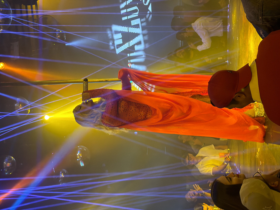 A blonde performer wearing a red sequined dress and flowing fabric hangs from a pole amidst blue laser lights and disco balls. She appears to be singing into a microphone. Below, a crowd of onlookers watches, some with raised hands. A large illuminated sign is visible in the background. Another individual wearing a red cap and jacket stands nearby. The scene is brightly lit with yellow and orange hues.