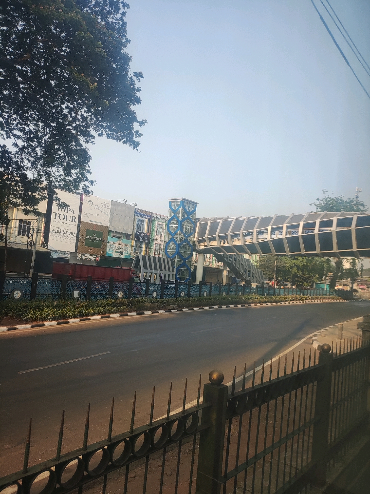 A pedestrian bridge spans a street in East Jakarta, Indonesia, during daylight. The bridge, with a modern, angular design, features a metal frame and dark, opaque panels. The street below is asphalt, with clear lane markings and a white and black striped curb. On the left side of the street, a row of buildings with advertisements is visible, including a large banner for "WIPA TOUR." A lush green tree with dark foliage overhangs the buildings. A black metal fence with pointed spikes runs along the foreground, separating the sidewalk from the street. The sky is a pale blue, indicating clear weather.