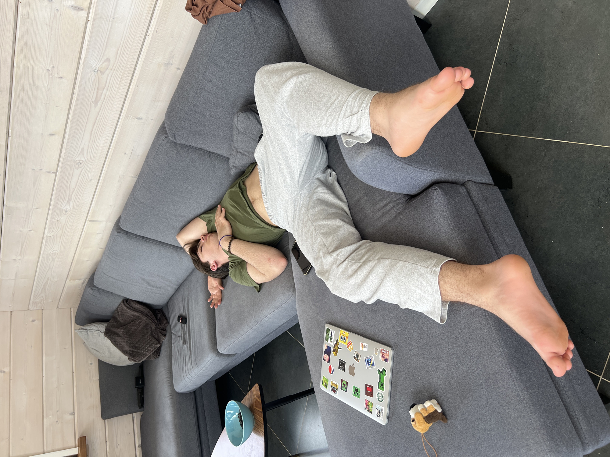 A young man is lying diagonally across a gray sectional sofa. He is wearing a green t-shirt and gray sweatpants. His arms are positioned near his head, seemingly resting or in a relaxed pose. A laptop covered in stickers is on the sofa near his feet. A small wooden toy is also next to the laptop. The floor is partially visible, showing a black tiled surface. The adjacent wall and ceiling are lined with white wooden planks. A small side table with a blue bowl on it is also visible next to the sofa. The city of Svetloe, Russia, is not identifiable in this image.